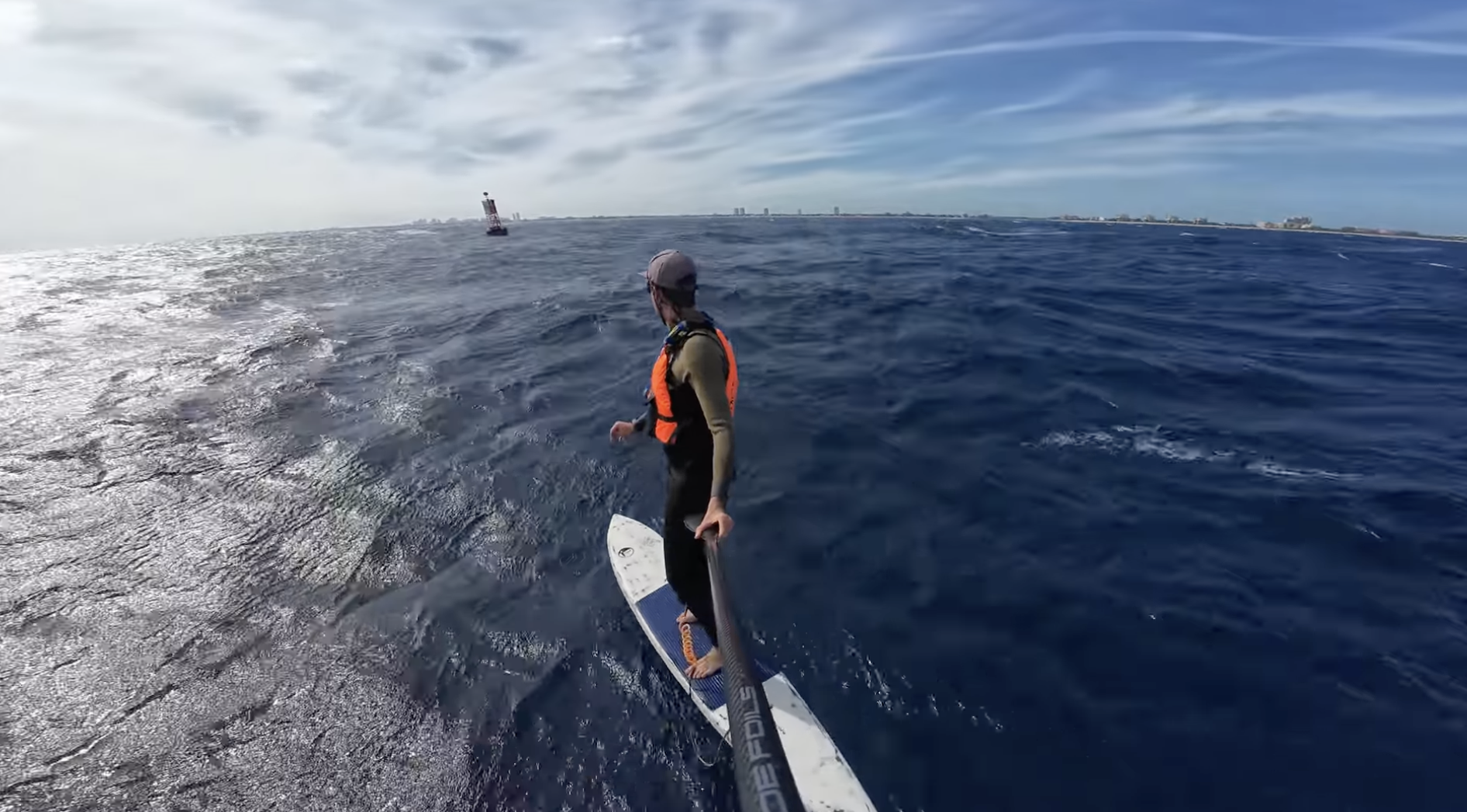 Nick Reeser riding a hydrofoil down winding on the open water, wearing a hat and a life vest, with a distant lighthouse and city skyline under a partly cloudy sky.