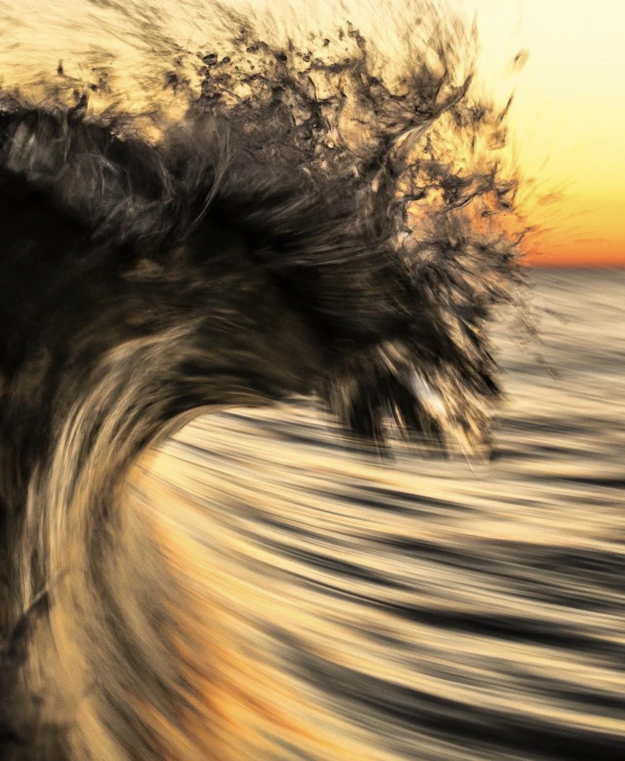 A large wave crashing with dark water spray against a sunset sky.