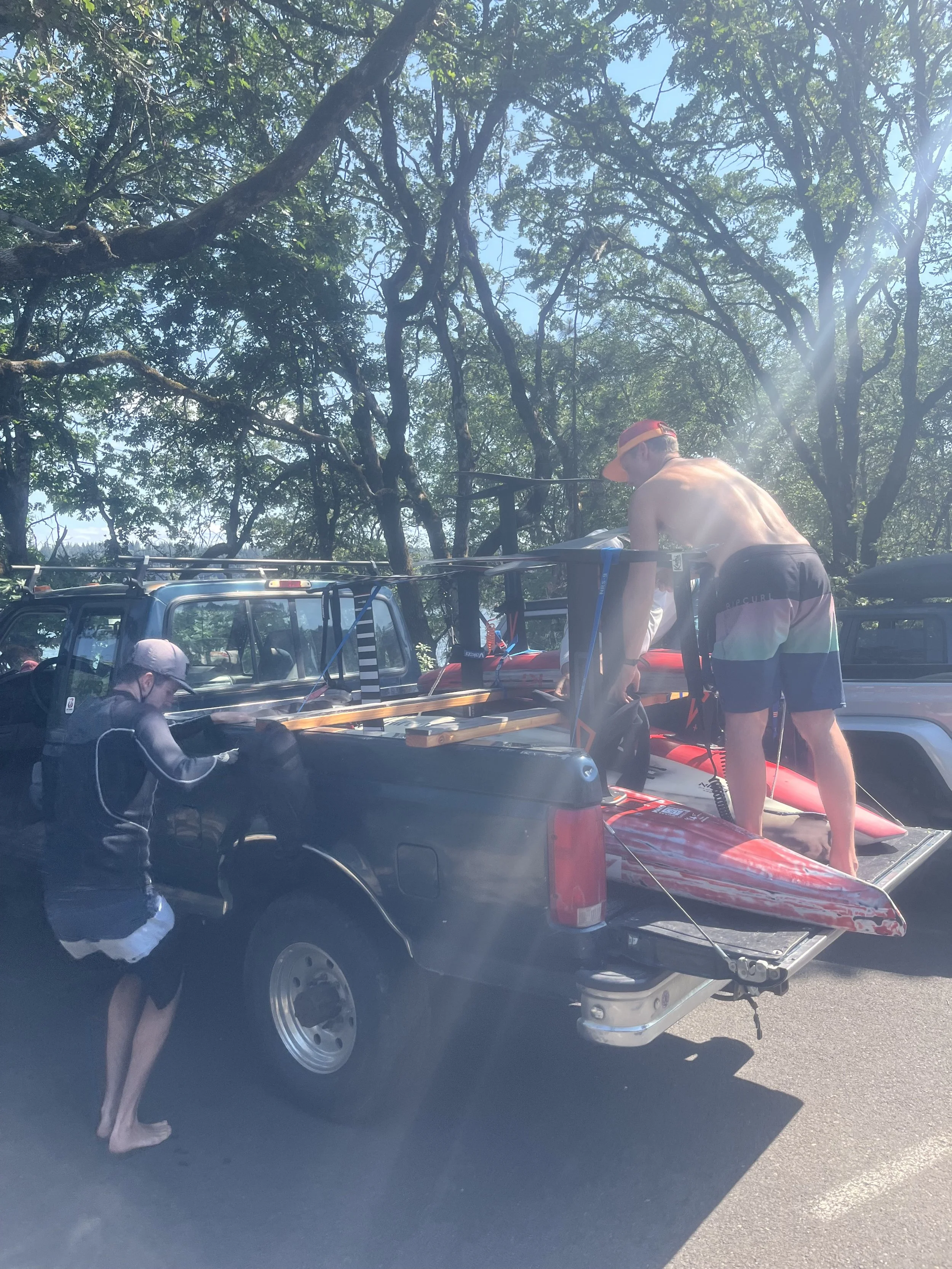 down wind shuttle with hydro foils in the back of a pickup truck, while another person is nearby wearing a cap and black clothing. There are trees and a clear blue sky in the background.