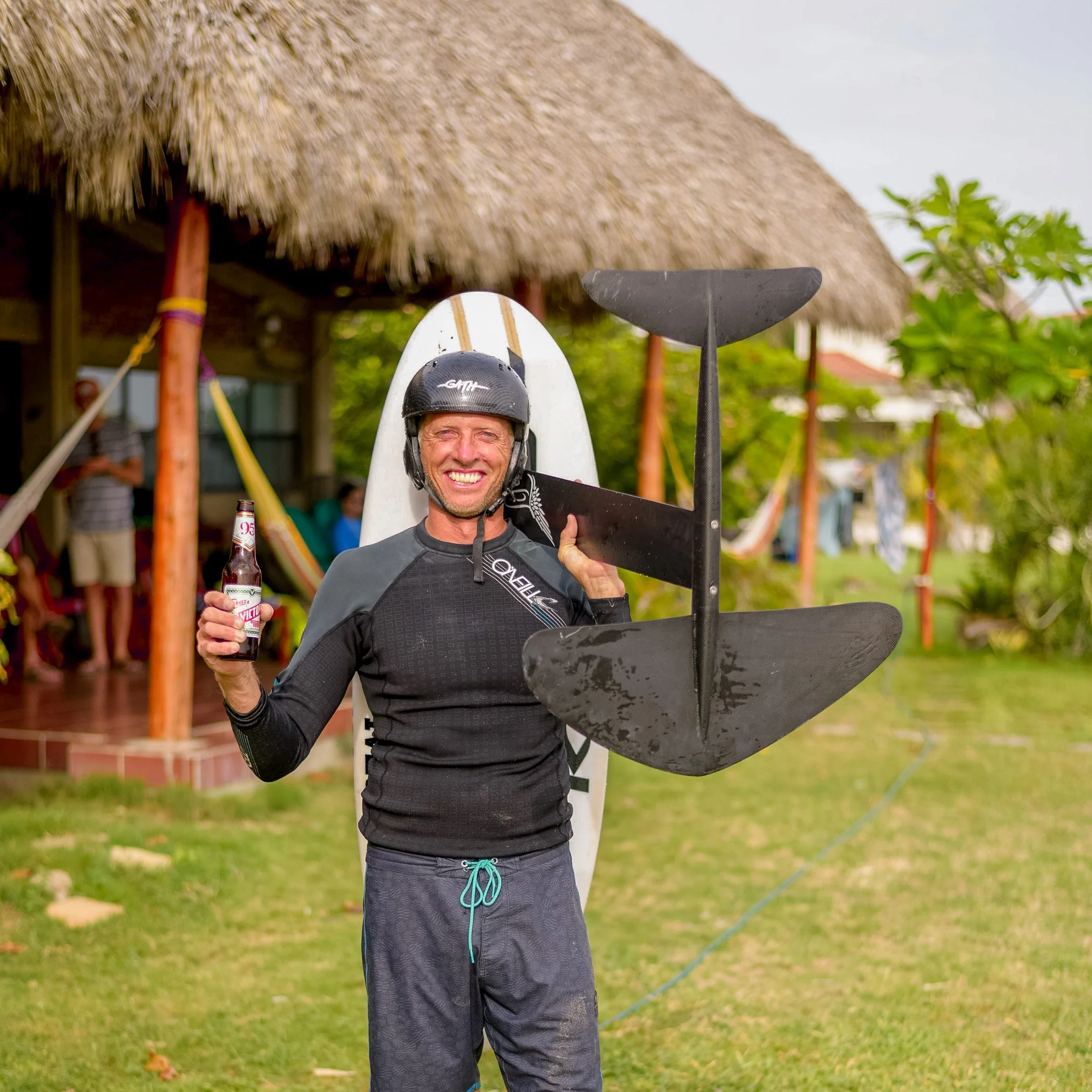 Seth Stafford smiling while holding a hydrofoil, wearing a helmet, in a tropical setting with a thatched hut and lush greenery in the background.