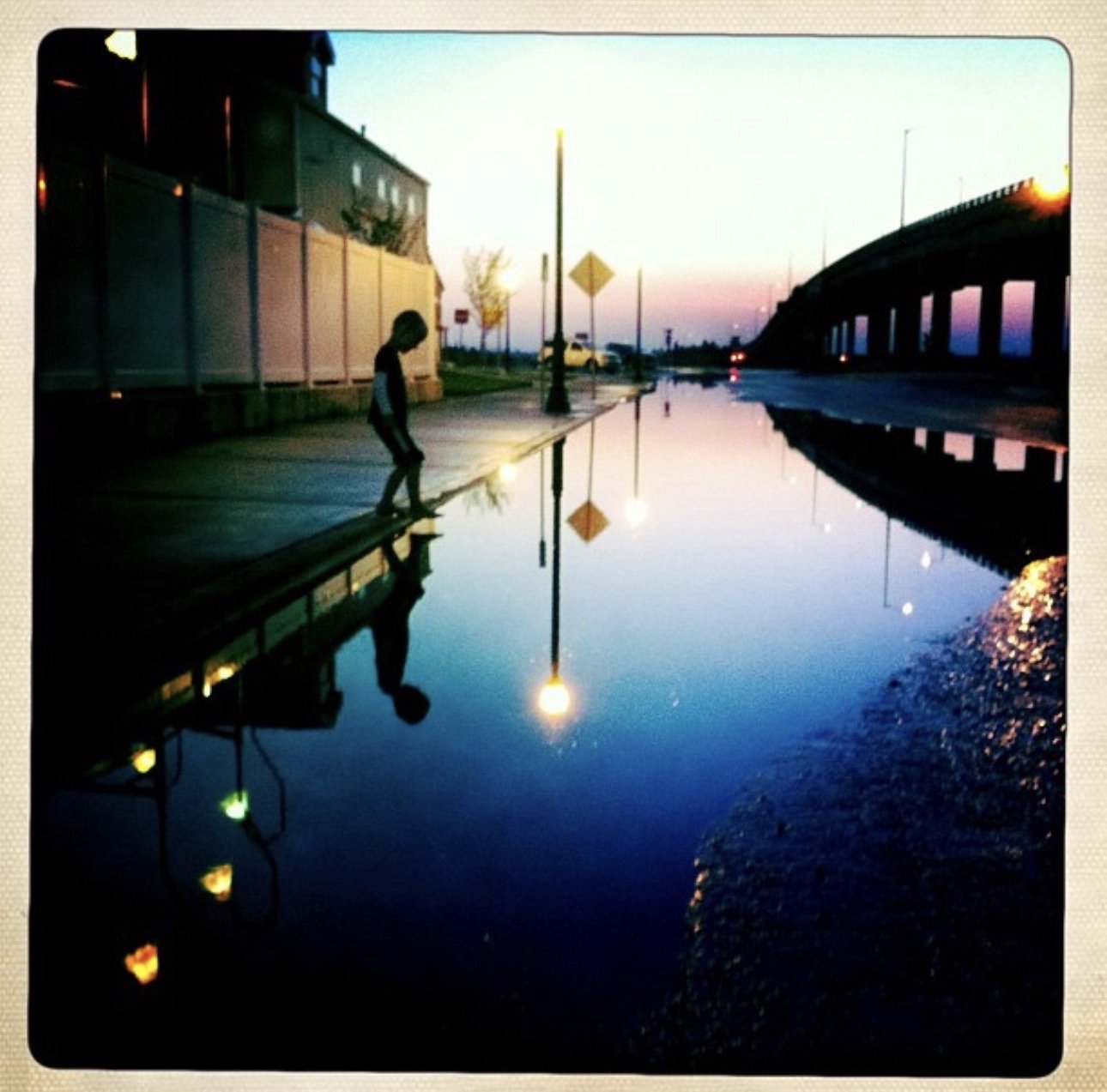 A boy walking along a sidewalk beside a large puddle reflecting the streetlights and sky at sunset.