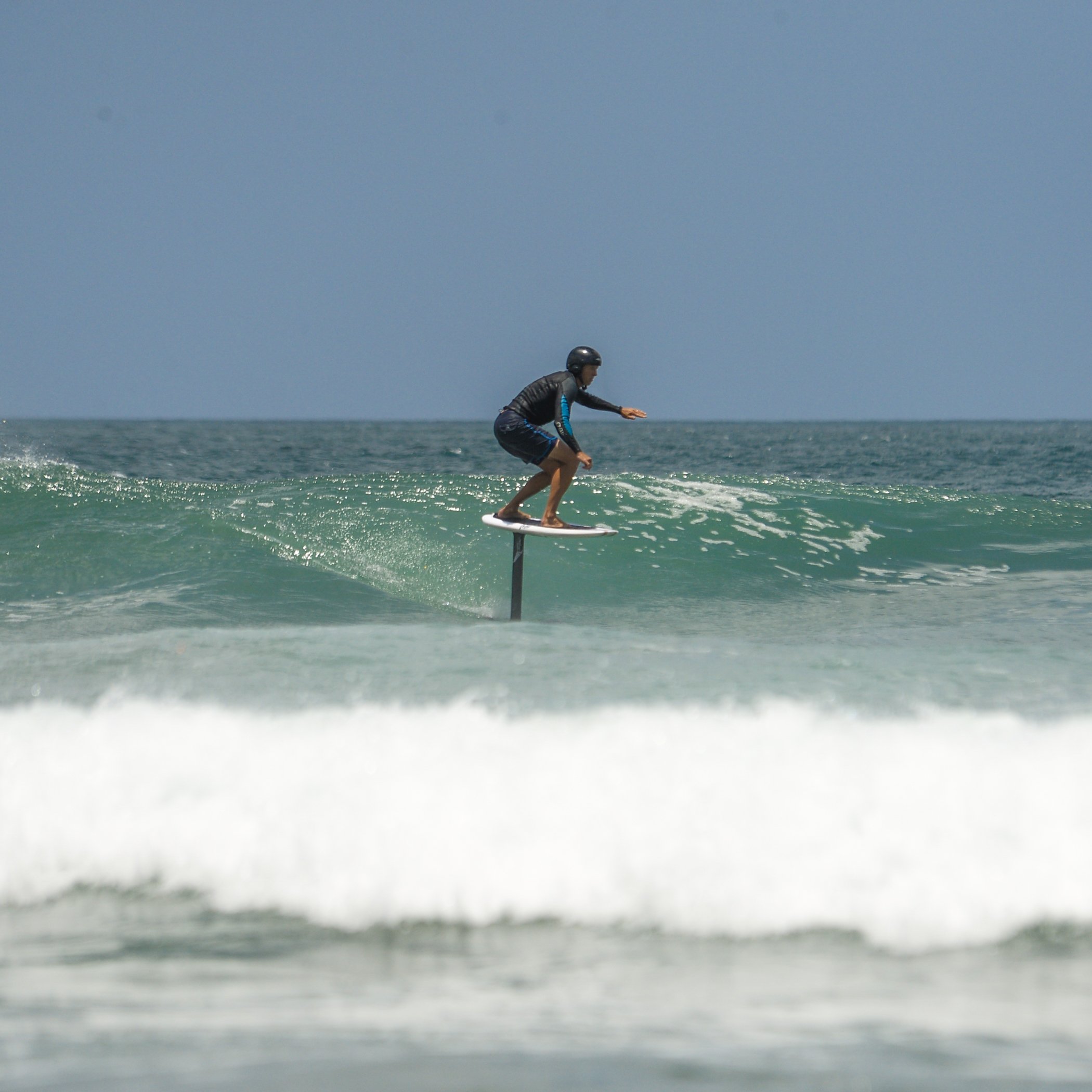 Seth Stafford wearing a helmet and wetsuit riding a hydrofoil board over ocean waves under a clear blue sky.