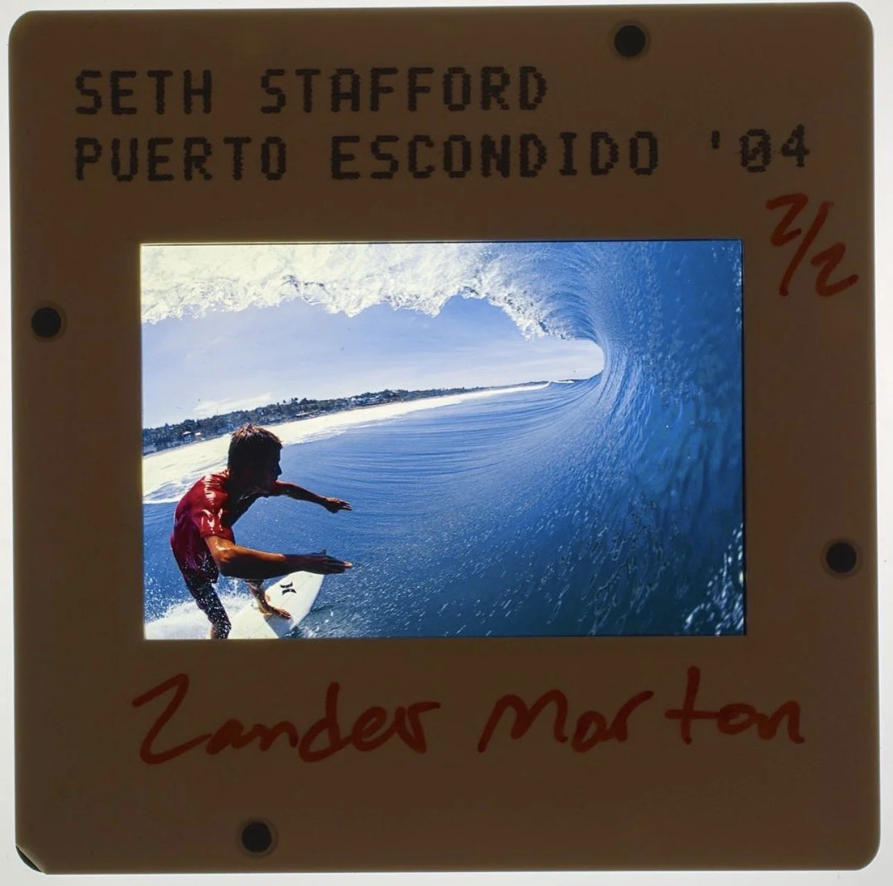 A framed photograph of a surfer riding a large blue wave, with the shoreline and houses in the background. The frame has handwritten text: "SETH STAFFORD PUERTO ESCONDIDO '04" at the top and "Zander Morton" at the bottom.