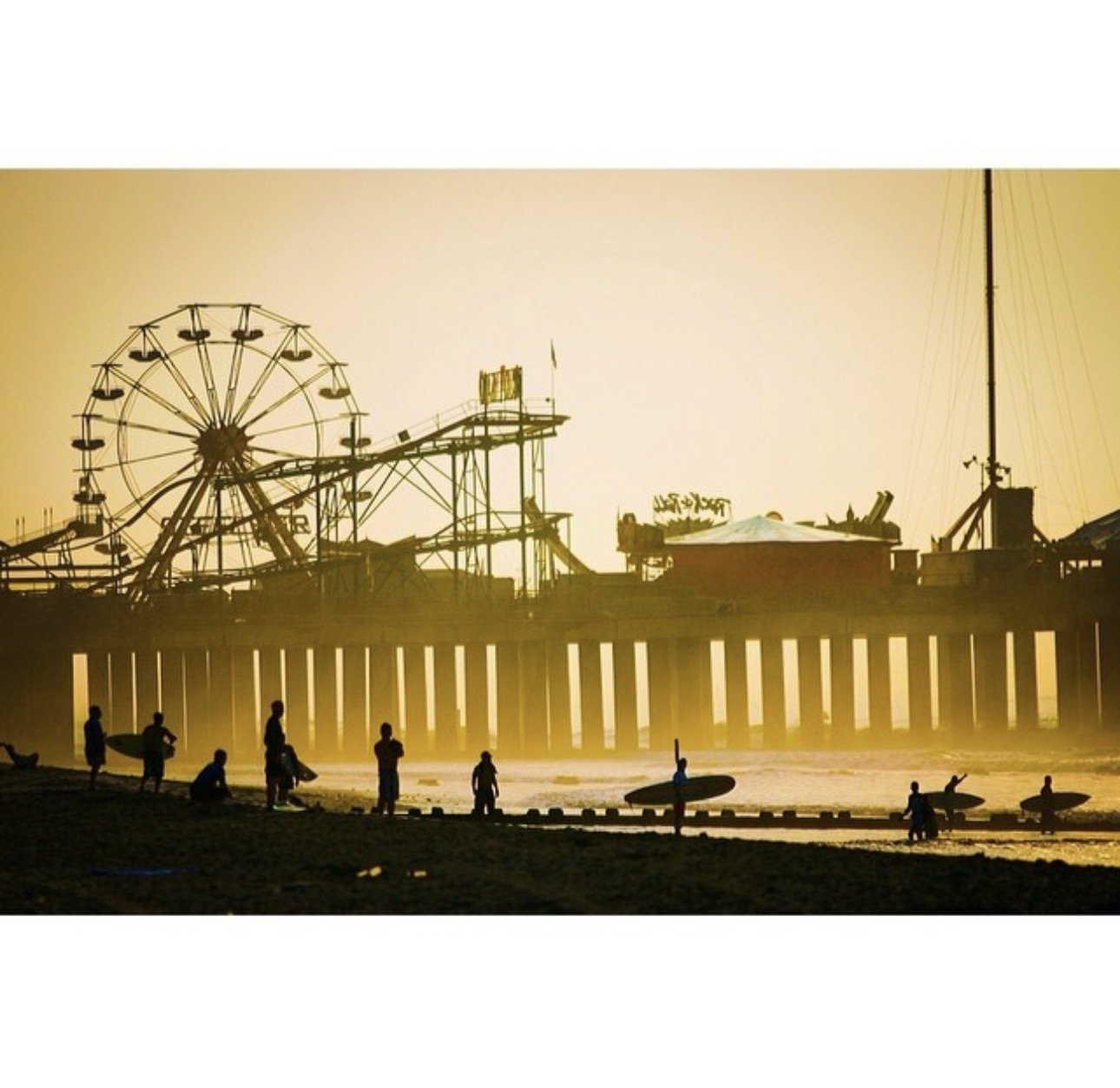 Silhouettes of people with surfboards on a beach at sunset, with an amusement park pier featuring a Ferris wheel and roller coaster in the background photographed by Seth Stafford.
