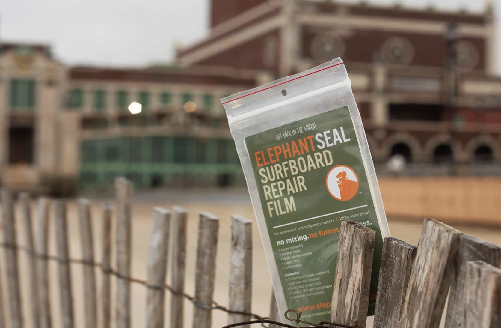 A bag of Elephant Seal Surfboard Repair Film in a wooden fence on a beach with buildings in the background.
