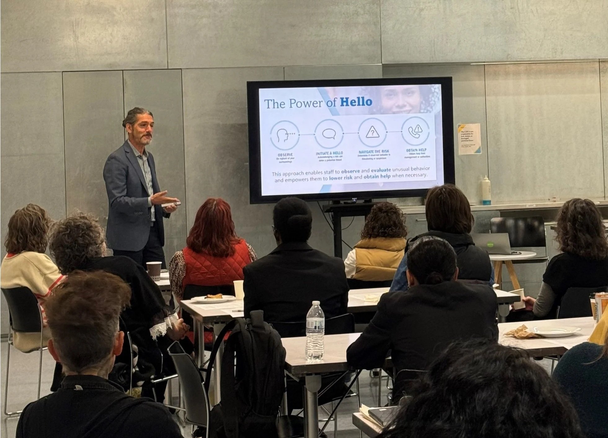 A man in a suit giving a presentation to an audience seated at tables in a conference room. A large screen behind him displays a slide titled 'The Power of Hello', with icons and text explaining the steps of a process related to greeting or communication. The room has gray walls, and several audience members are visible, some with laptops and notebooks, listening attentively.