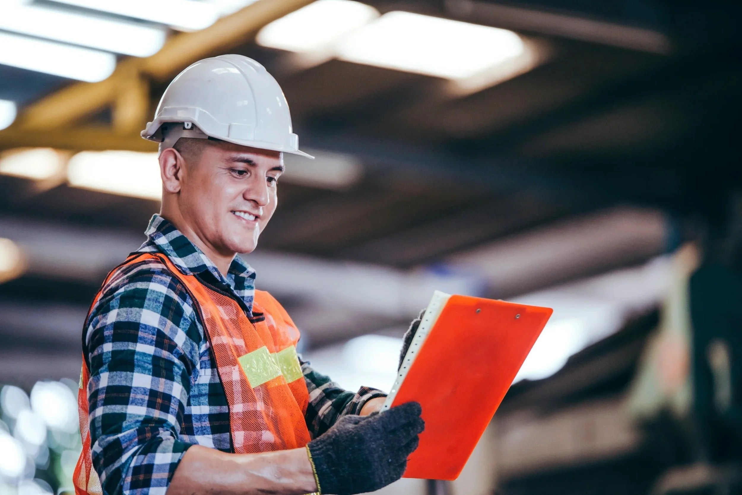 A young male construction worker in a plaid shirt, orange safety vest, white hard hat, and black gloves holding an orange clipboard, smiling and looking at it in a warehouse or industrial setting.