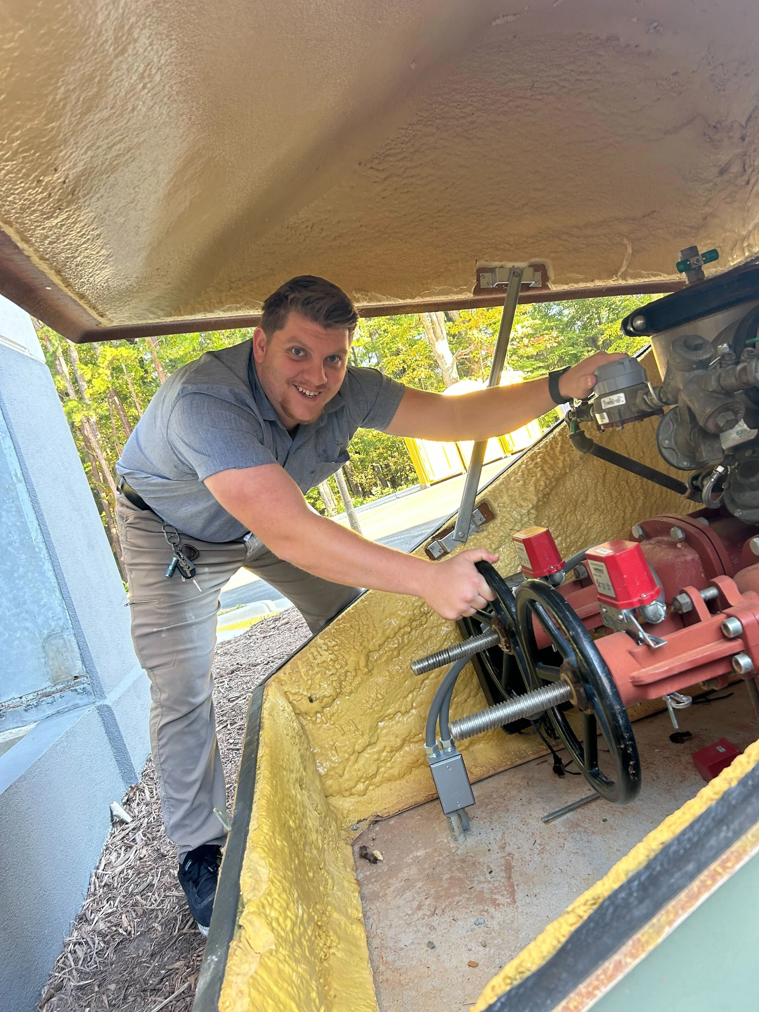 A man with short brown hair wearing a gray shirt and beige pants inspecting and working with mechanical equipment inside a hollowed yellow foam structure outside. Behind him, green trees and a blue building are visible.