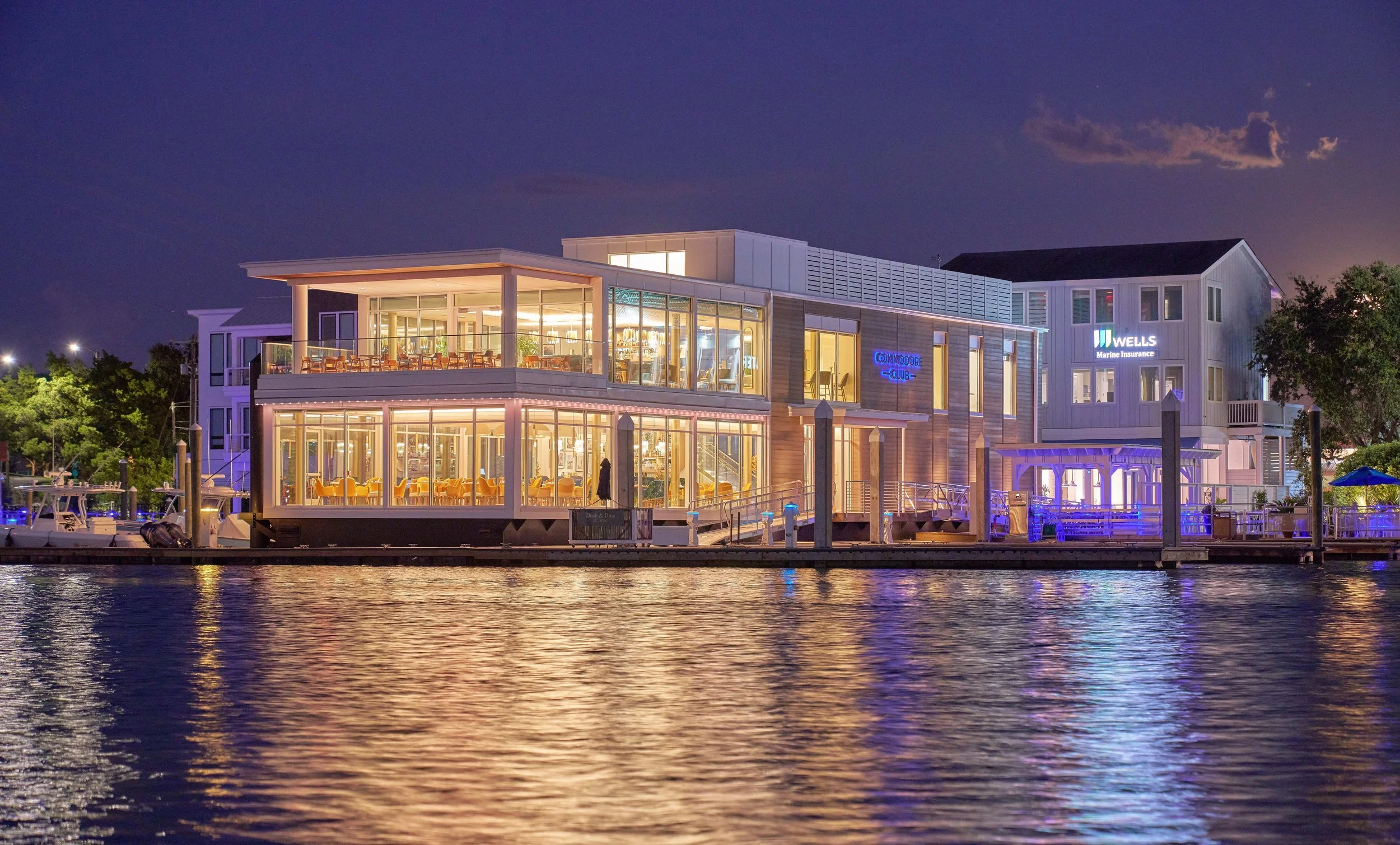 A modern waterfront building at night with large glass windows, illuminated interior, and signs for 'Commodore Club' and 'Wells Marine Insurance'; boats are docked along the water in the foreground and trees are visible in the background.