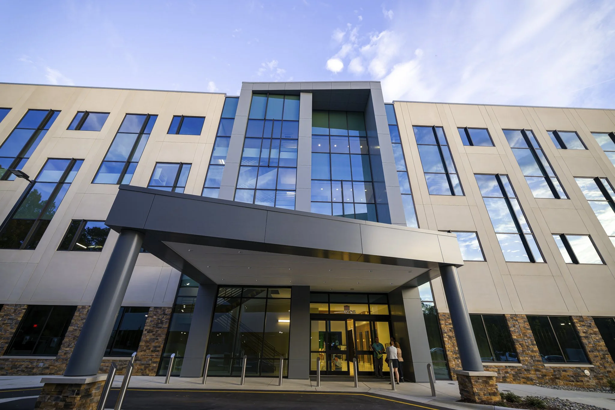 Modern commercial building with large glass windows, entrance with glass doors, and people standing at the entrance on a paved area under a partly cloudy sky.