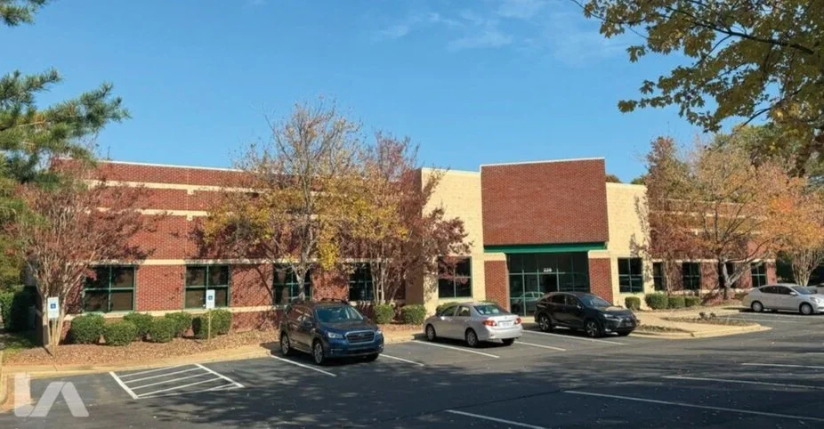A brick commercial building with several windows, surrounded by trees with fall foliage, and a parking lot with a few parked cars.