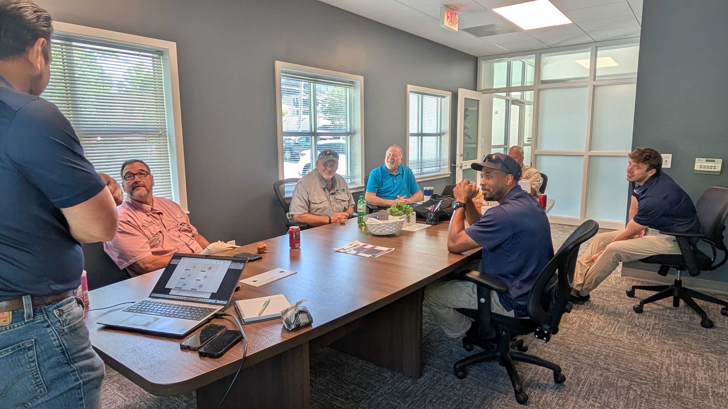 A group of seven men gathered in a conference room, seated around a large table. Some are smiling and engaging, with laptops, drinks, and notebooks on the table. The room has windows with blinds, and a glass door leads outside. An exit sign is visible on the ceiling.