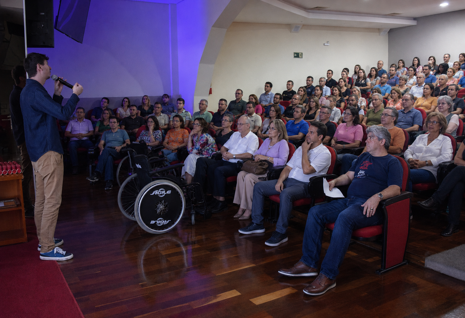 A man holding a microphone speaking to an audience seated in a lecture hall. The audience is paying attention, with some taking notes. The hall has red chairs and wooden flooring, and there is a wheelchair in front near the speaker.