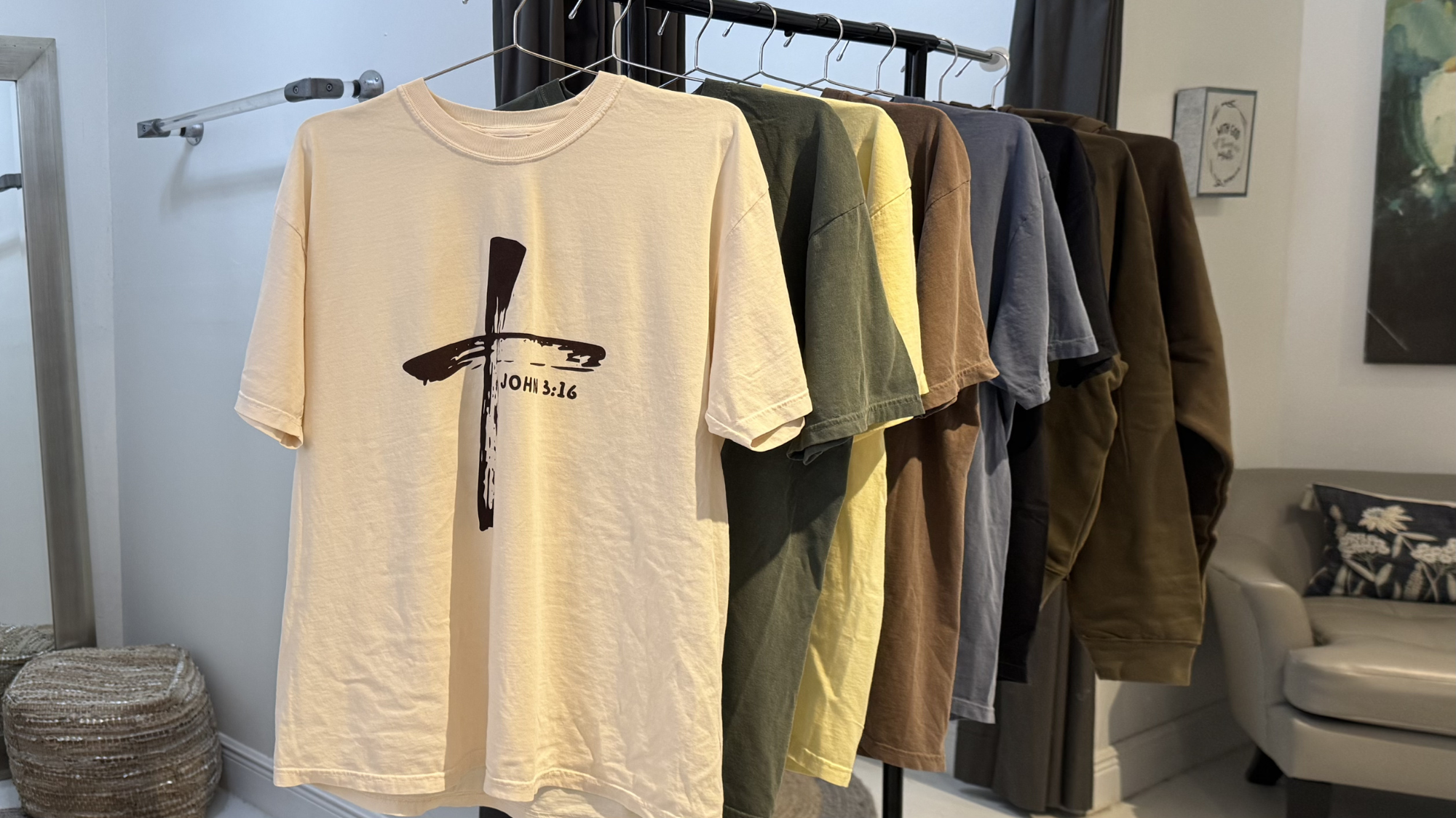 A clothing rack with six T-shirts in various colors including beige, green, yellow, blue, and brown. The beige T-shirt in front has a black cross with the Bible verse John 3:16 printed on it.
