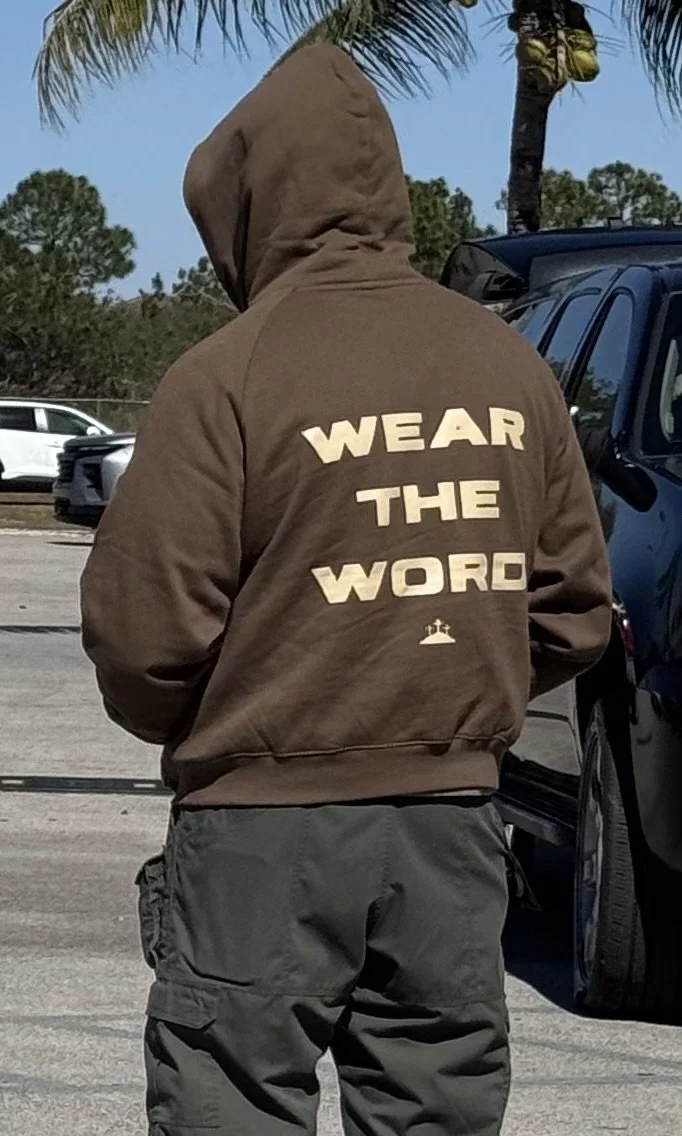 Person wearing a brown hoodie with the words "Wear the Word" and a small cross symbol on the back, standing in a parking lot with trees and blue sky in the background.