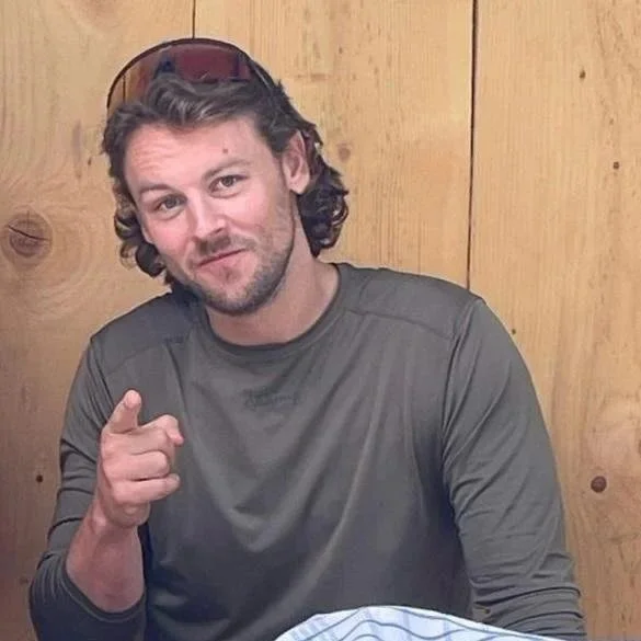 A man with dark, curly hair and a beard, wearing a gray long-sleeve shirt, sitting in front of a wooden wall, pointing at the camera with a slight smile.