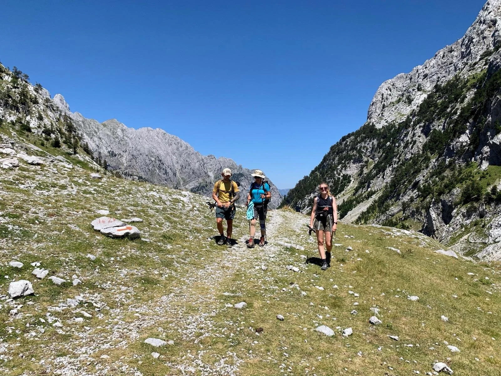 Three hikers walking on a trail in a mountainous landscape with rocky slopes and green grass under a clear blue sky.