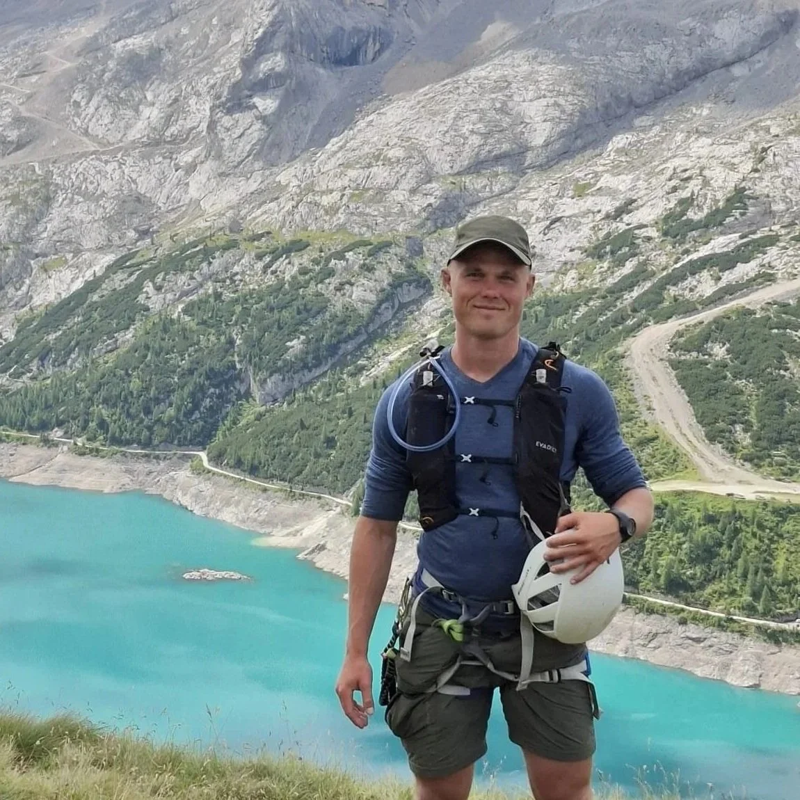 A man hiking near a blue lake with mountains in the background, holding a white helmet and wearing a backpack and outdoor gear.