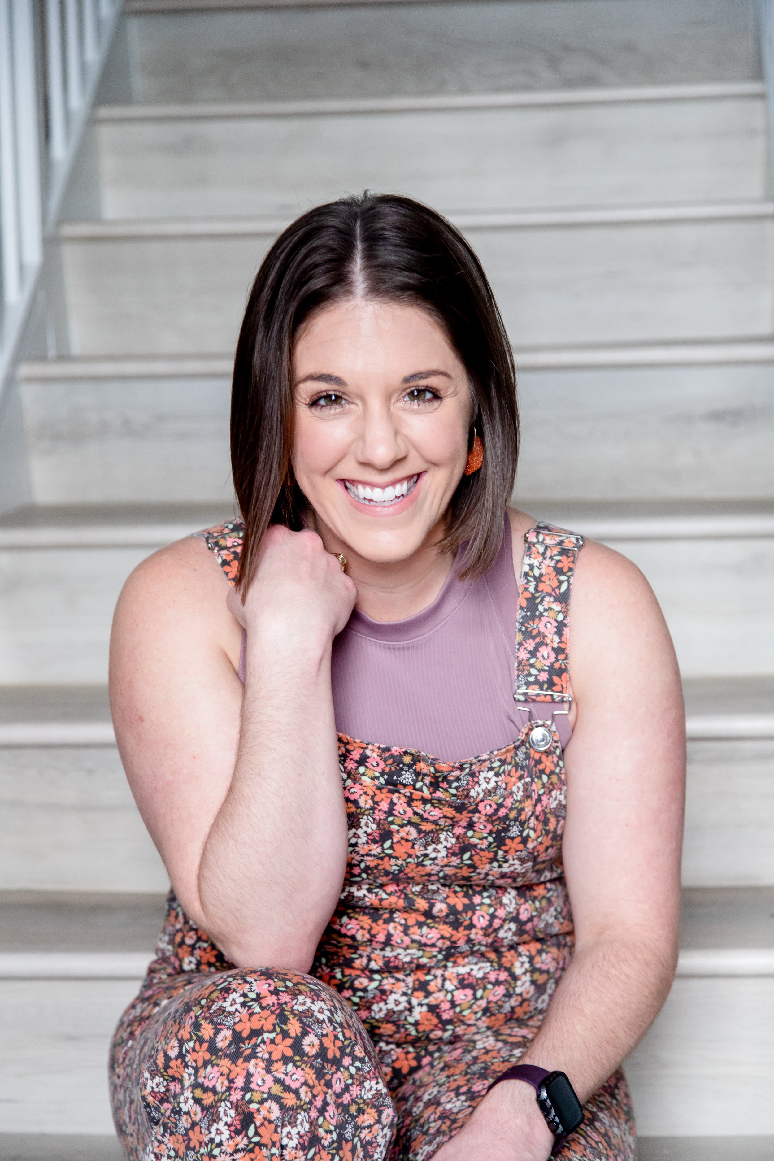 Business strategist AnnMarie Rose sitting on steps smiling, wearing a floral overall dress and a purple top