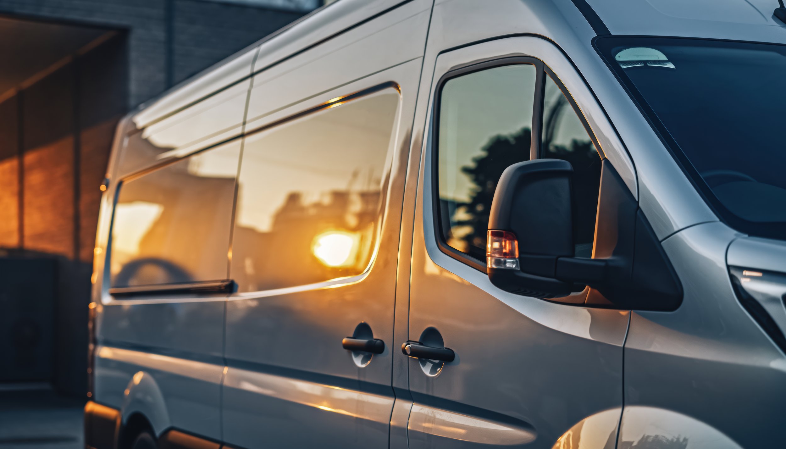 Close-up of a white delivery van during sunset, showing the front and side with a reflection of trees and sky on its windows.
