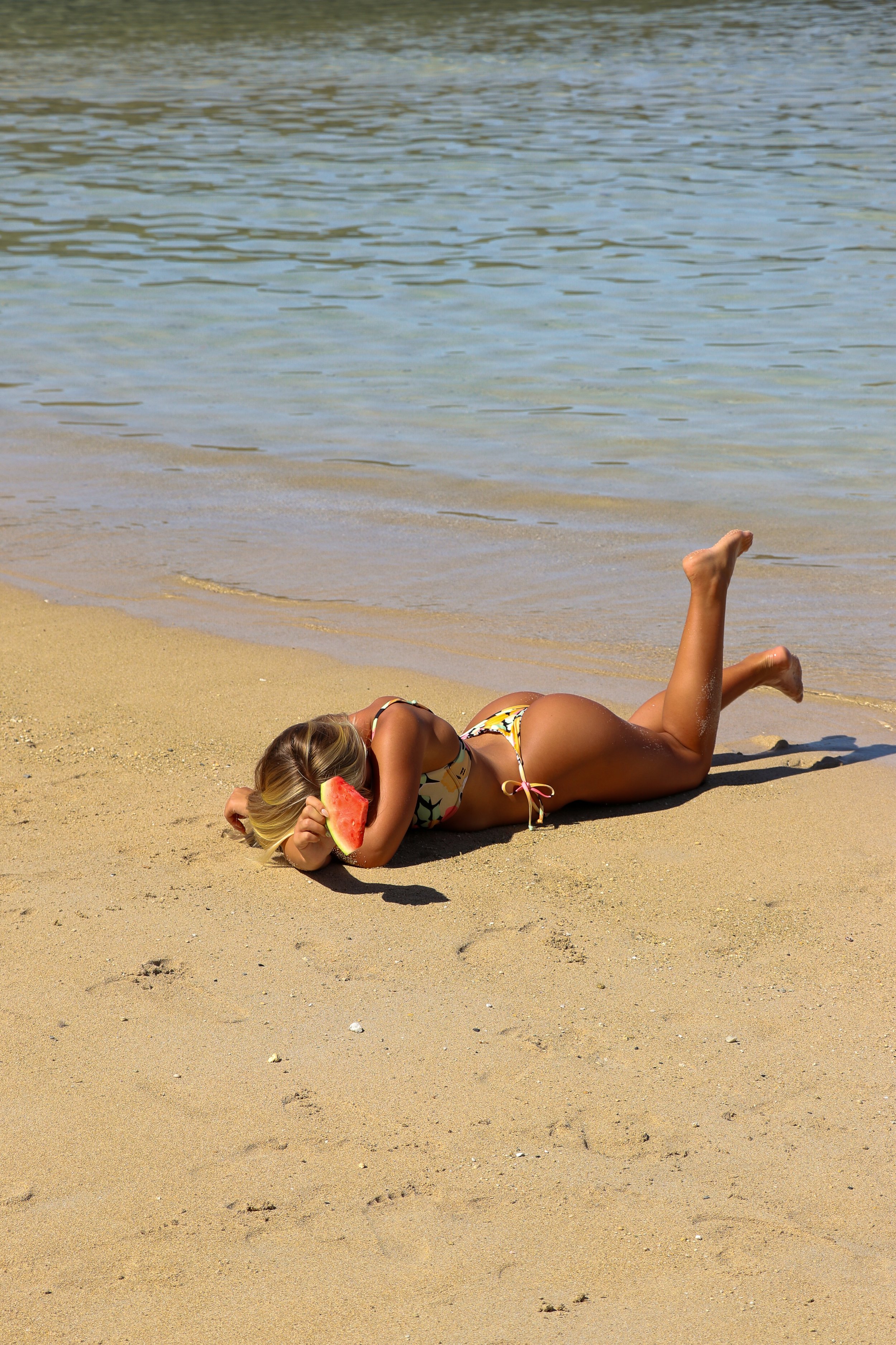 A woman lying on the sandy beach near the water, holding a slice of watermelon to her mouth.
