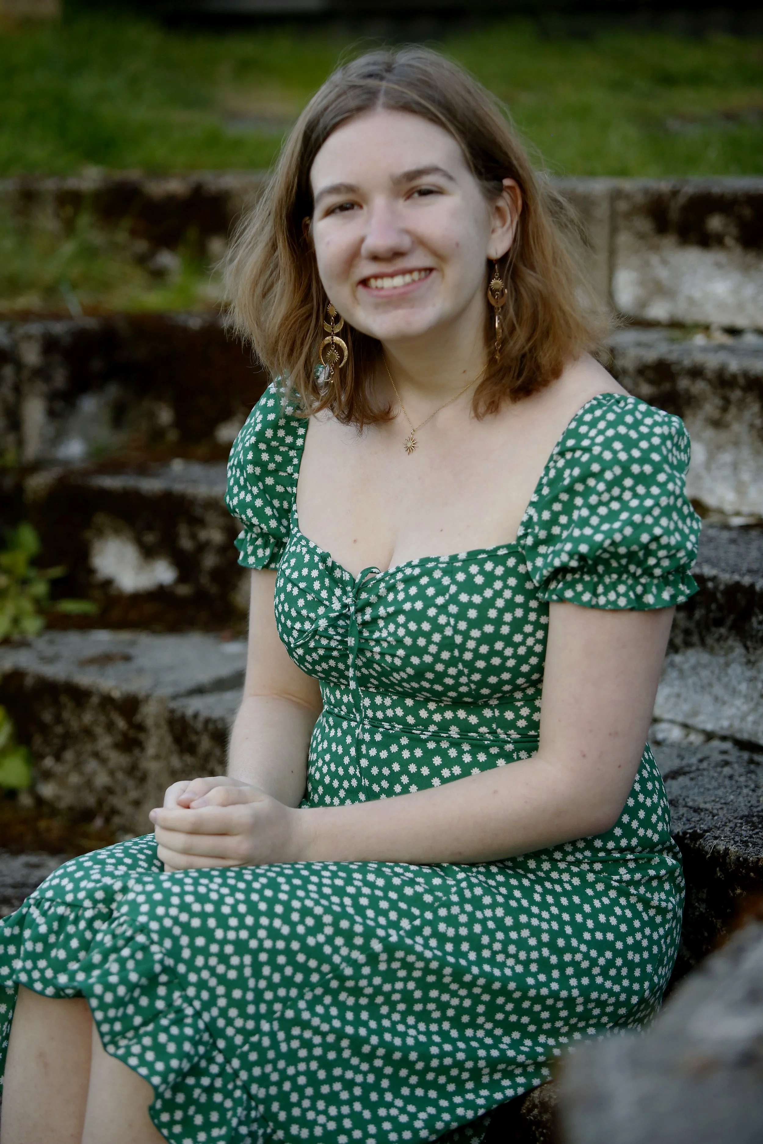 Smiling woman in green polka dot dress sitting outdoors near rocks with grass background.