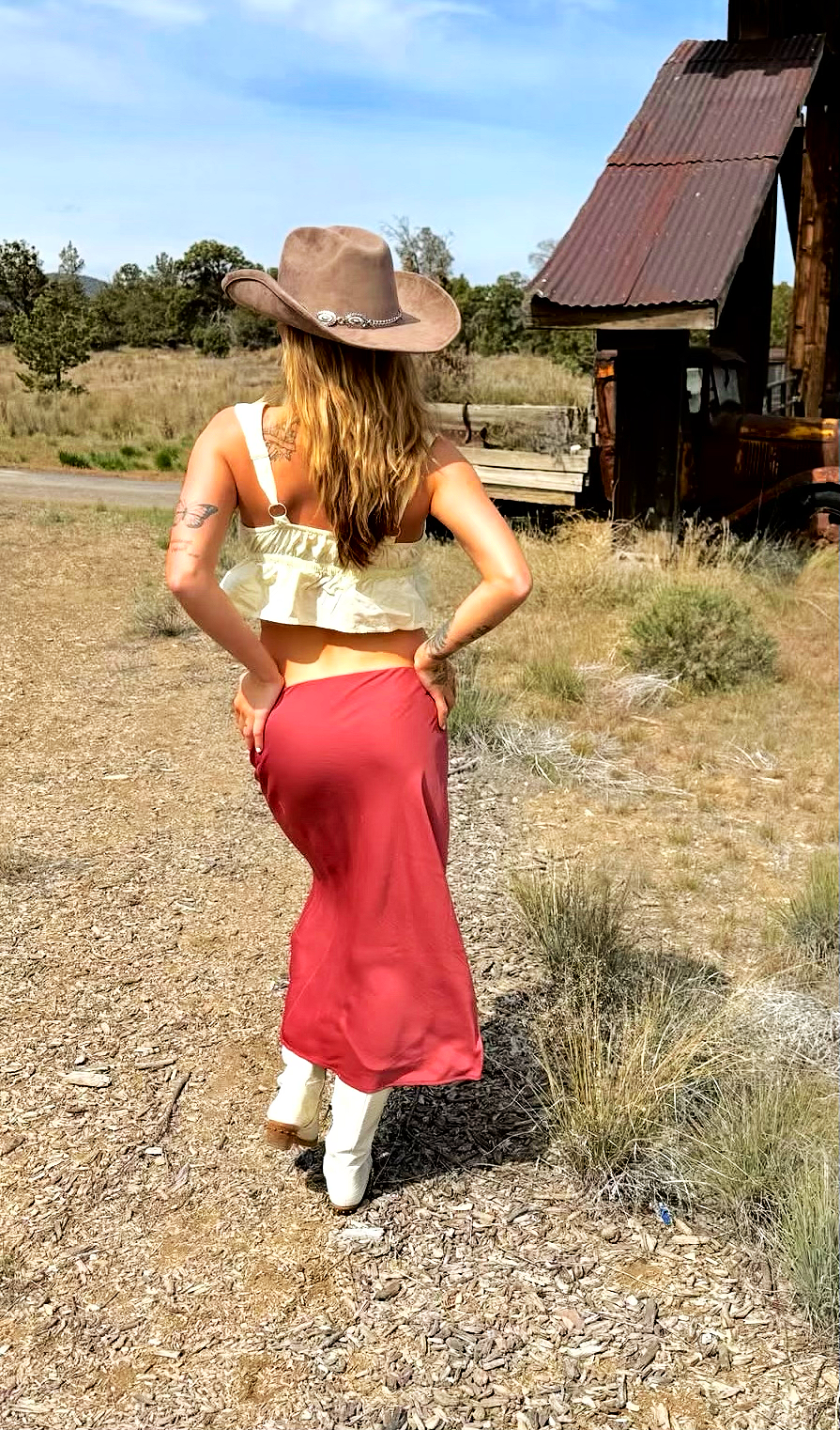 A woman with long wavy hair wearing a wide-brimmed cowboy hat, a cream-colored crop top, a long red skirt, and white boots, standing on a dirt path in a rural area with a rustic building in the background under a partly cloudy sky.
