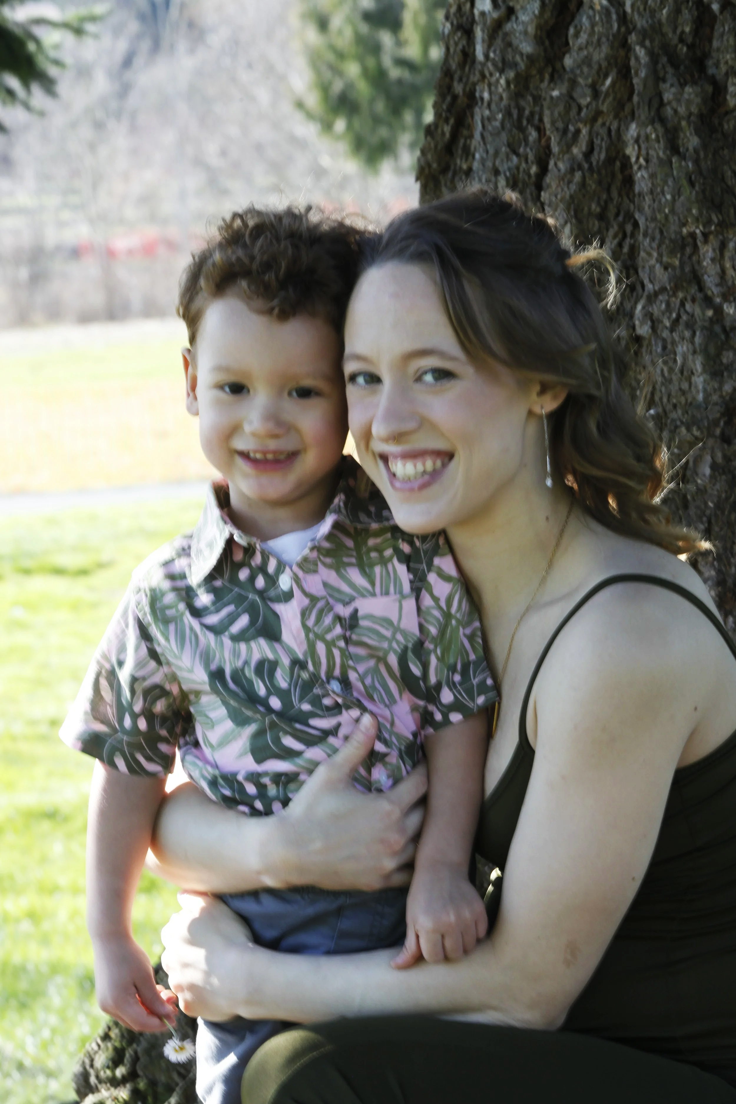 A woman and a young boy hugging outdoors near a tree, smiling at the camera with a park background.
