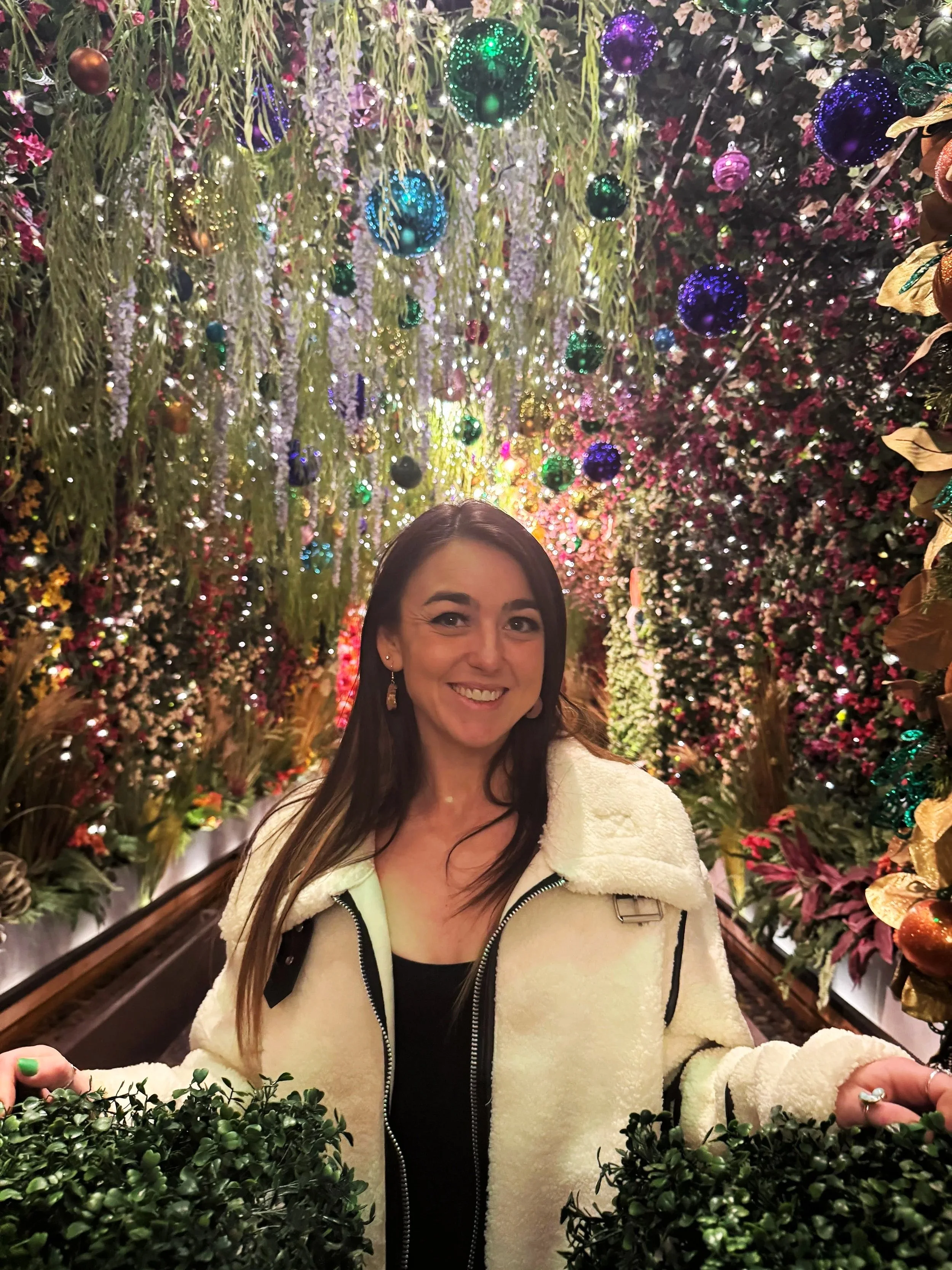 Woman smiling in a decorated, festive setting with colorful ornaments, flowers, and lights hanging from the ceiling.