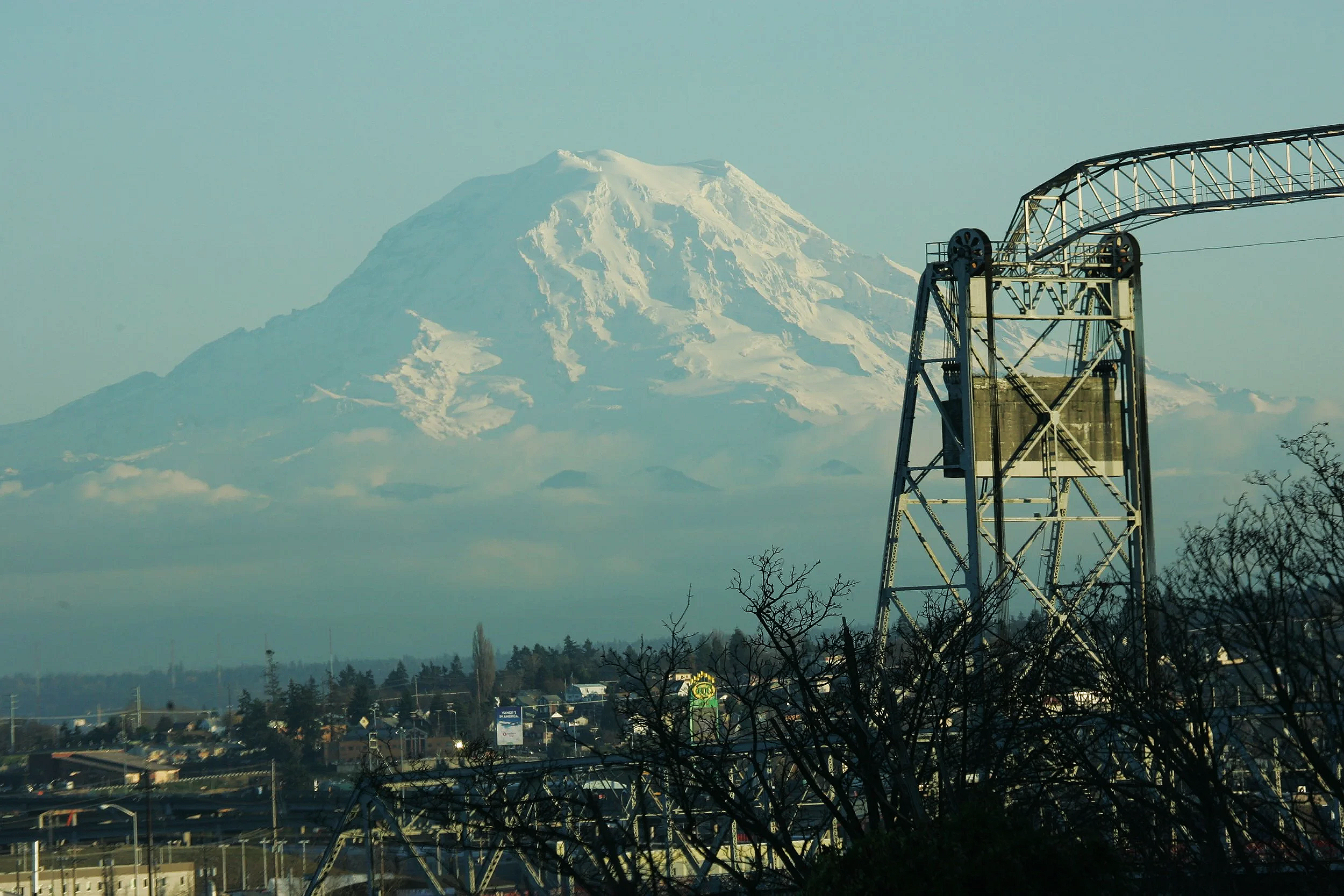 Snow-capped mountain in the background with an industrial structure and a cityscape in the foreground.