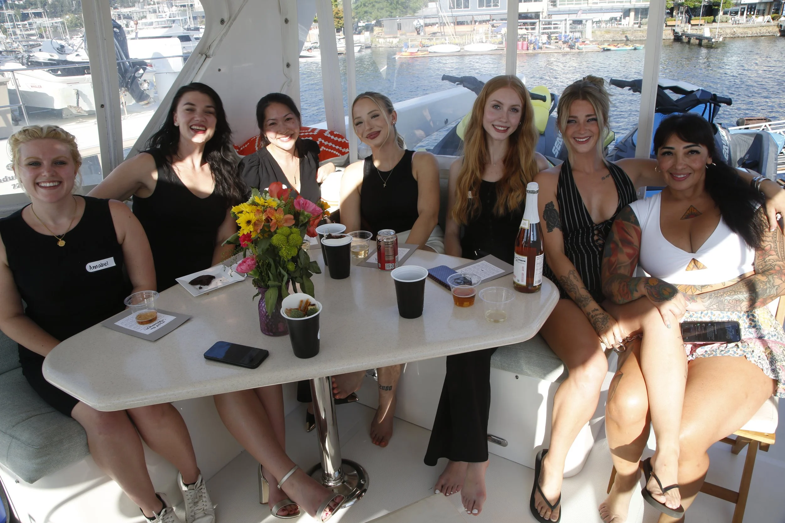 Seven women sitting at a table on a boat with a marina in the background, smiling and enjoying drinks and snacks.