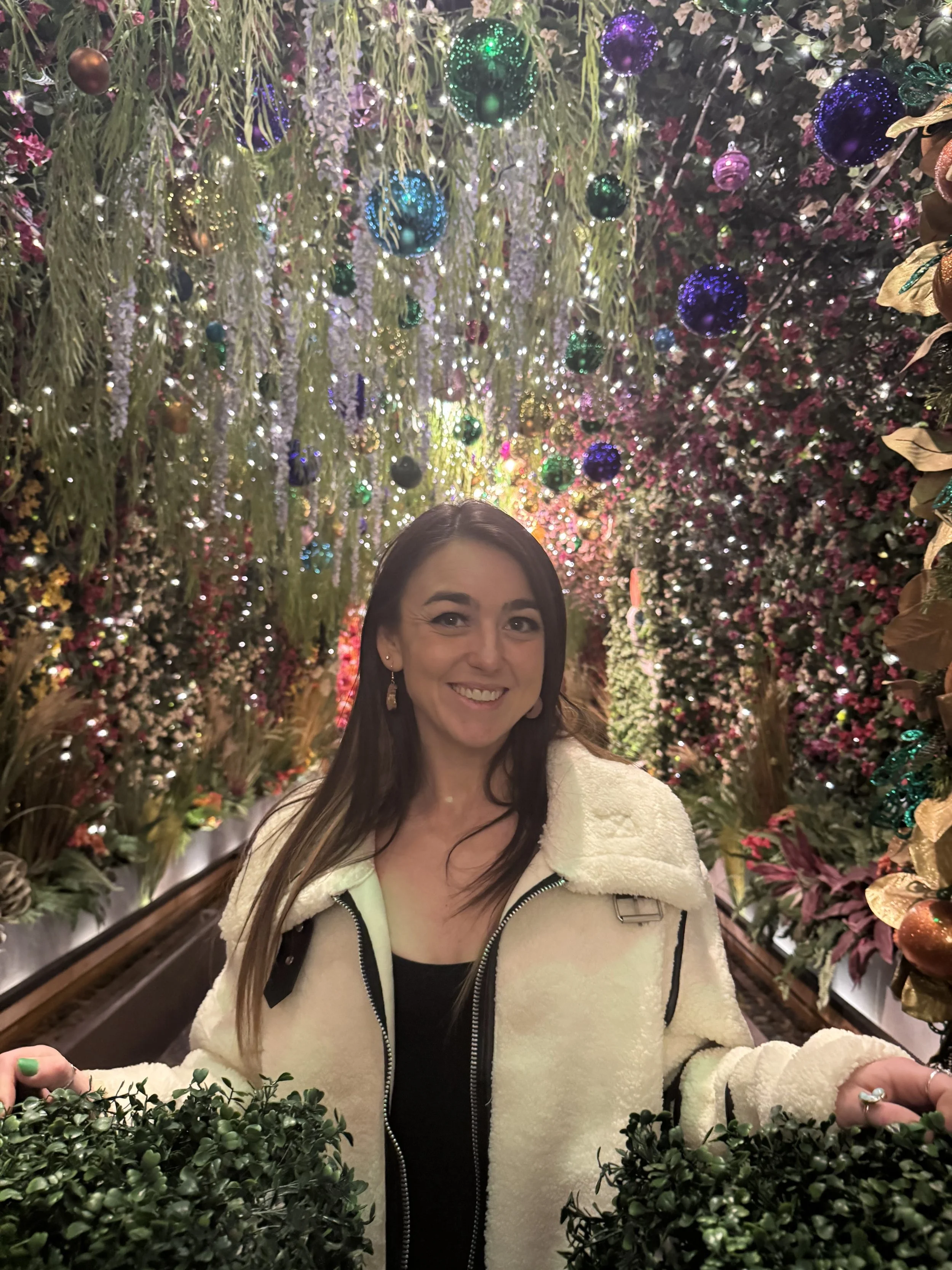 Woman smiling in a decorated, festive setting with colorful ornaments, flowers, and lights hanging from the ceiling.