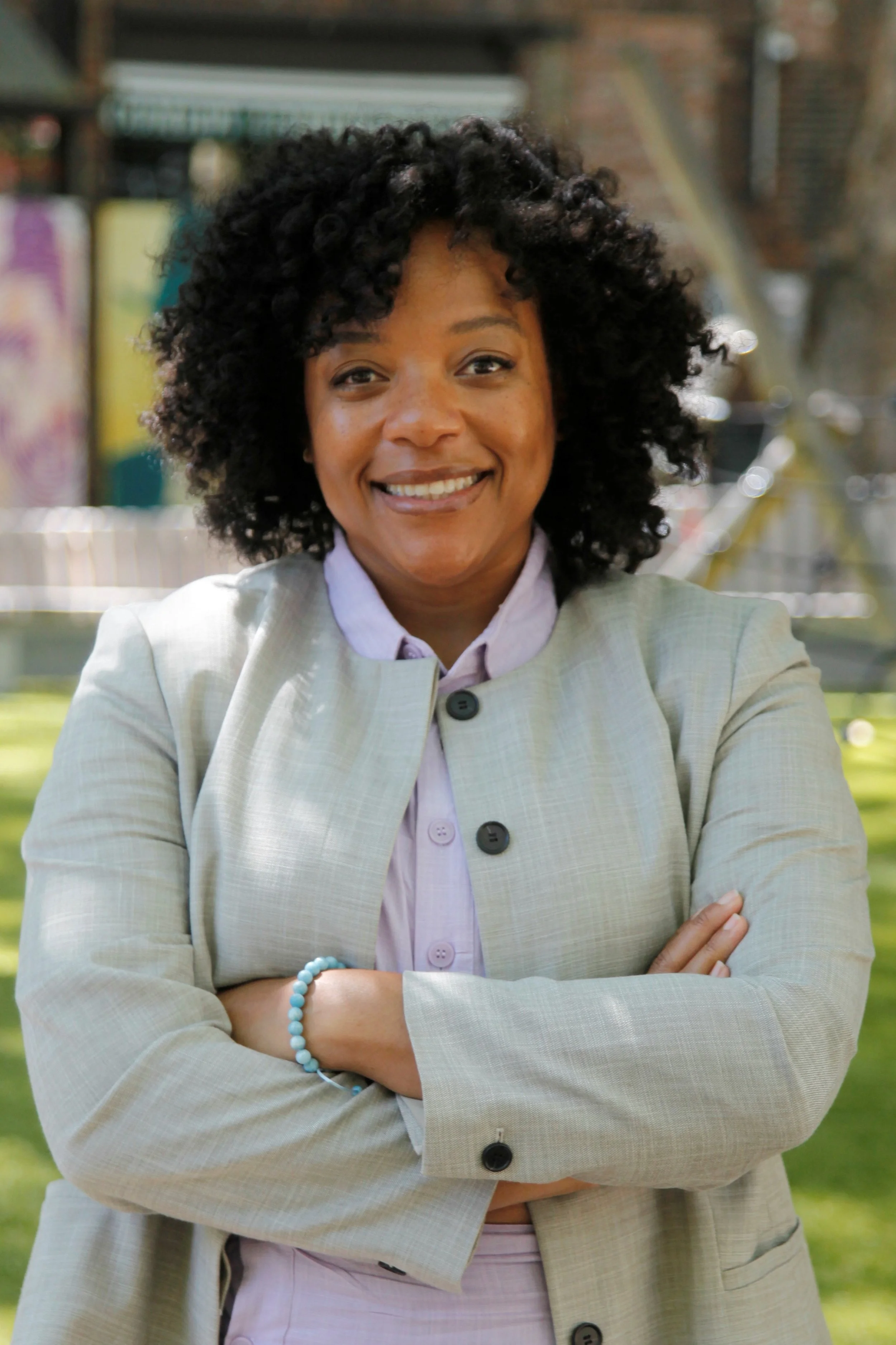 A woman with curly black hair smiling at the camera, wearing a light-colored blazer and a purple shirt underneath. She is standing outdoors with her arms crossed and a background of trees, a bicycle, and buildings.