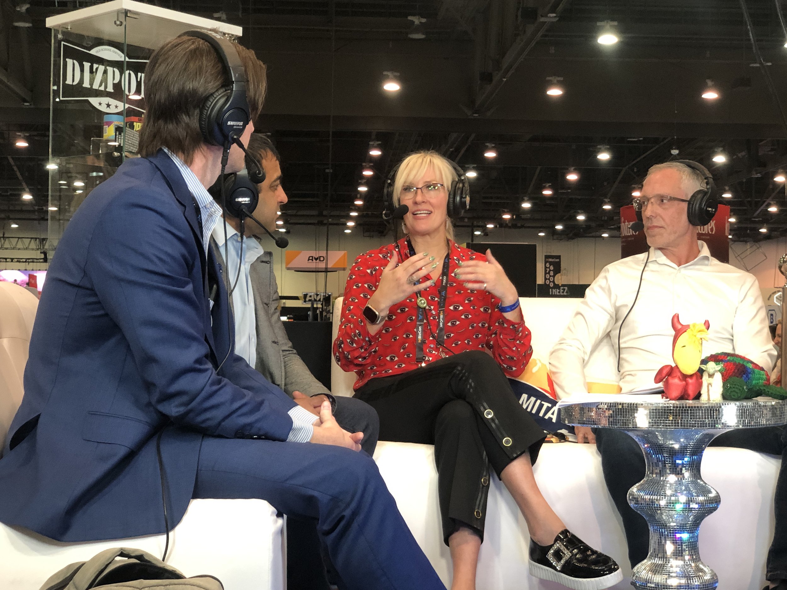 Four people sitting around a table wearing headphones with microphones, engaging in a conversation at what appears to be a conference or event, with a decorative rooster sculpture on the table.