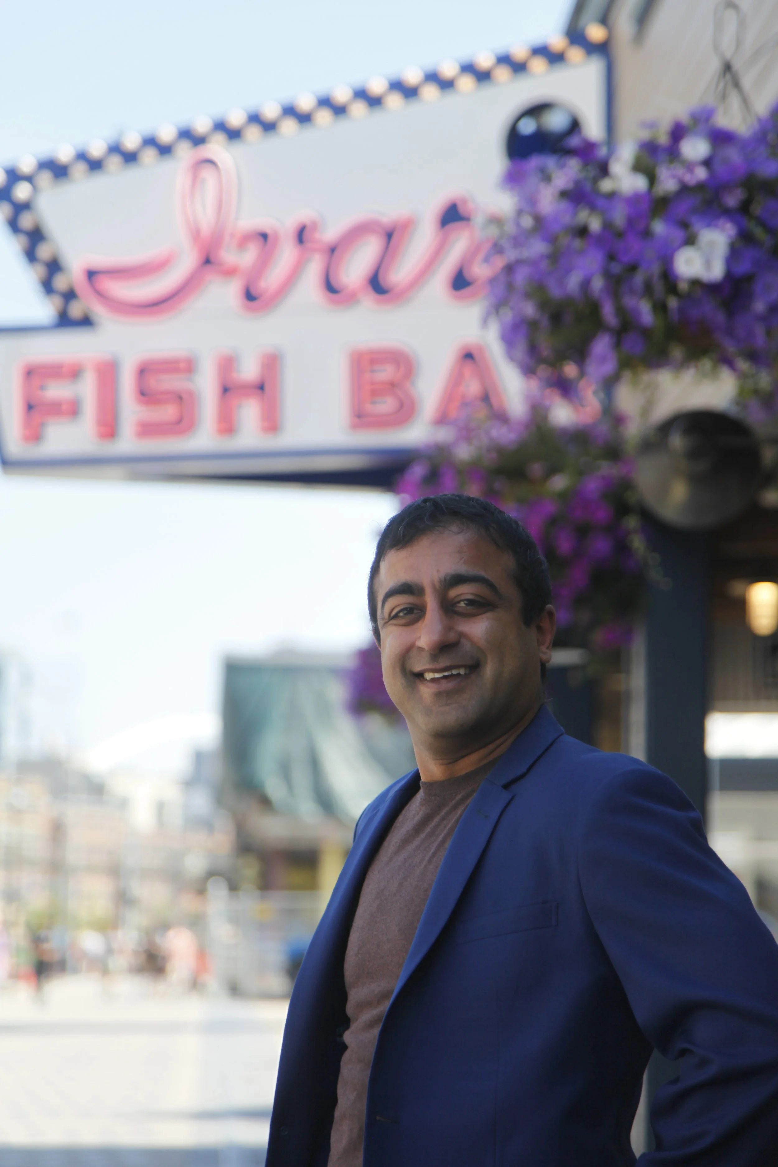 A man in a blue blazer smiling outdoors near a sign that reads 'Seafood Fish Bar' with purple flowers nearby.