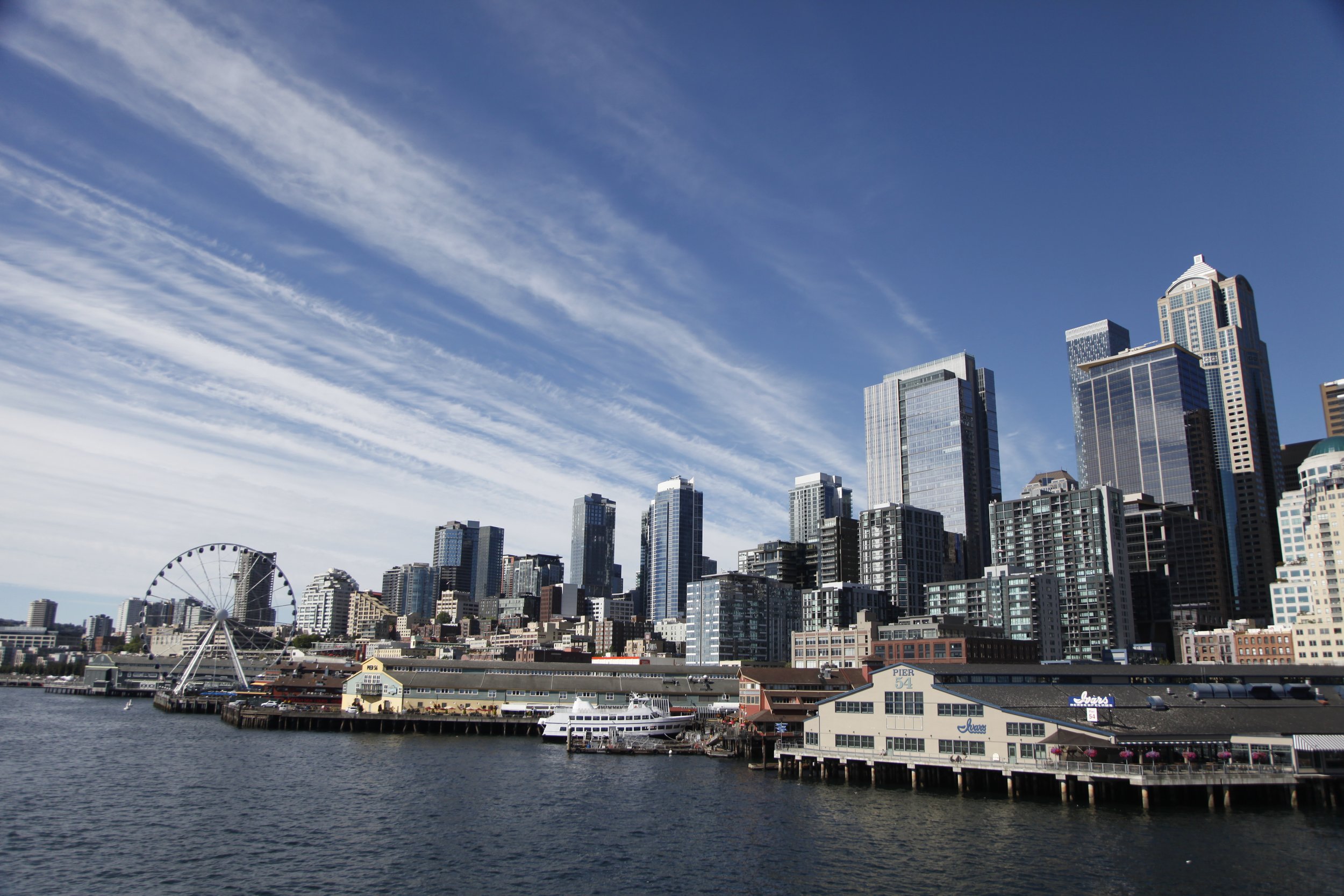Seattle waterfront with high-rise buildings, a Ferris wheel, and boats docked at the pier under a partly cloudy sky.