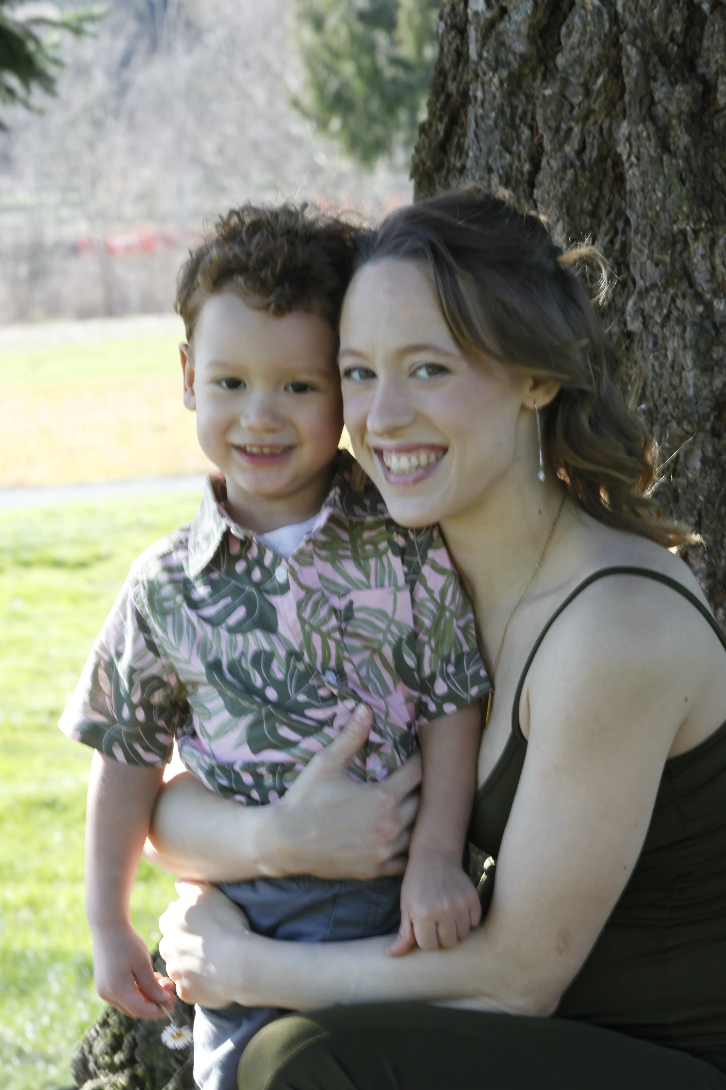 A woman and a young boy hugging outdoors near a tree, smiling at the camera with a park background.