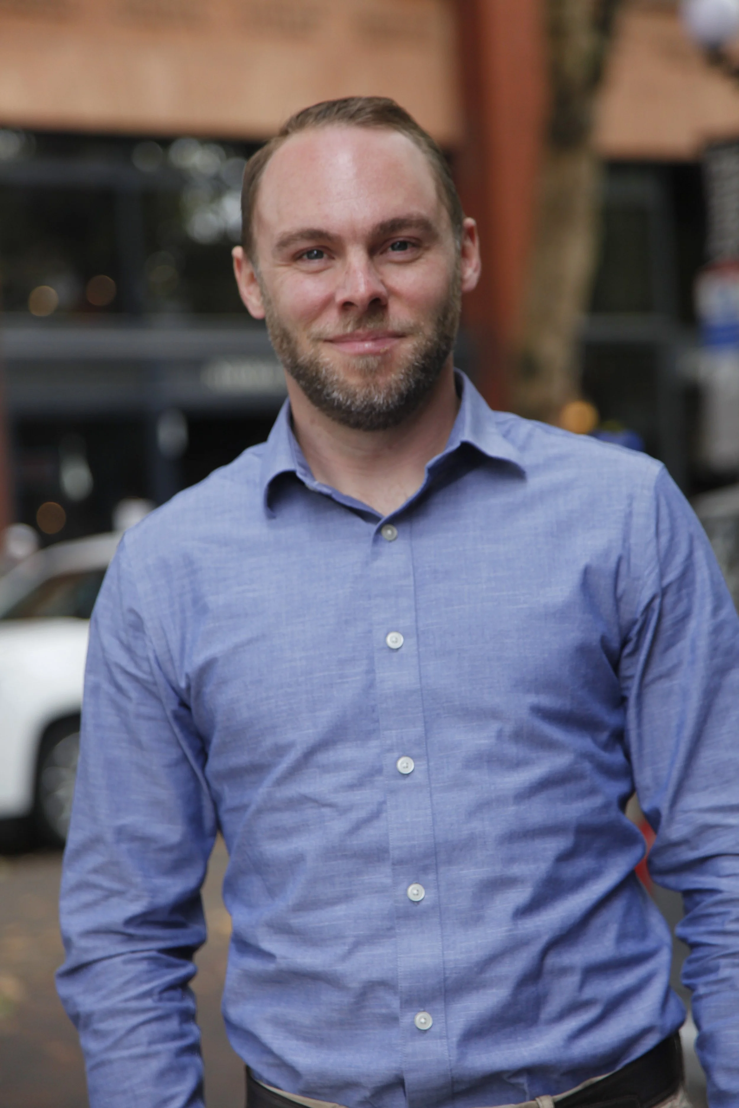 A smiling man with short, brown hair, wearing a blue button-down shirt, standing outdoors with a blurred background.