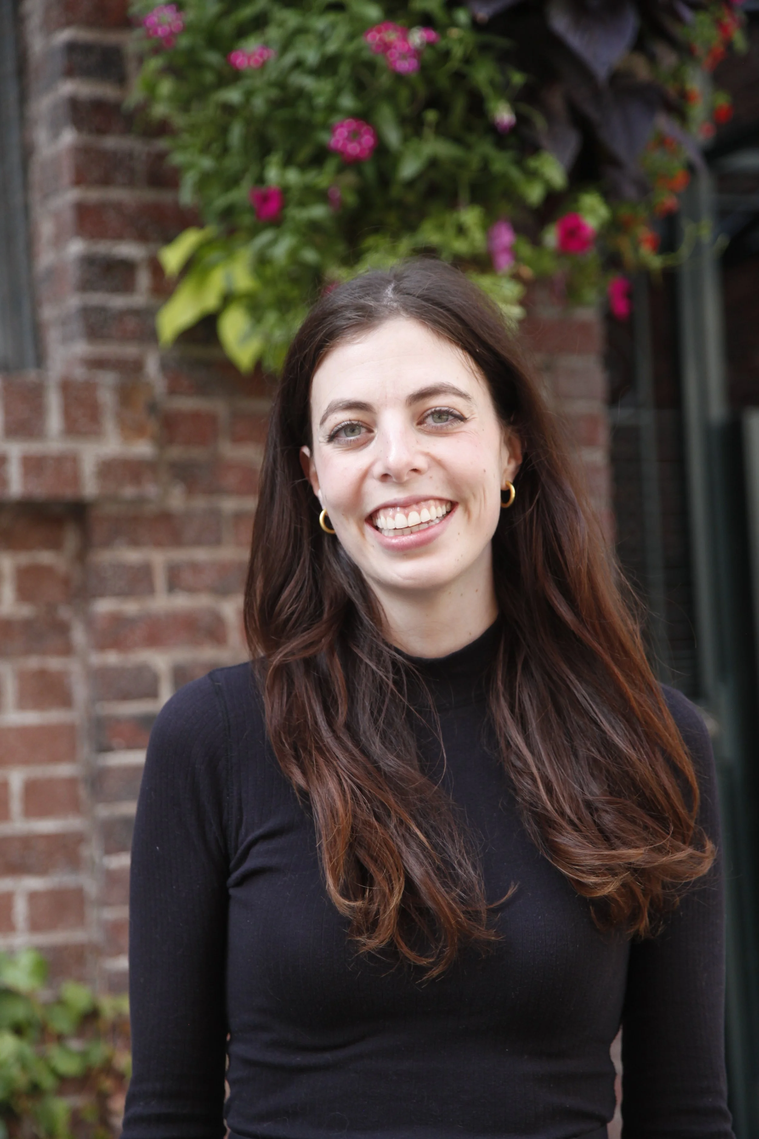 A young woman with long brown hair, wearing a black top and gold hoop earrings, smiling outdoors in front of a brick wall and blooming pink flowers.