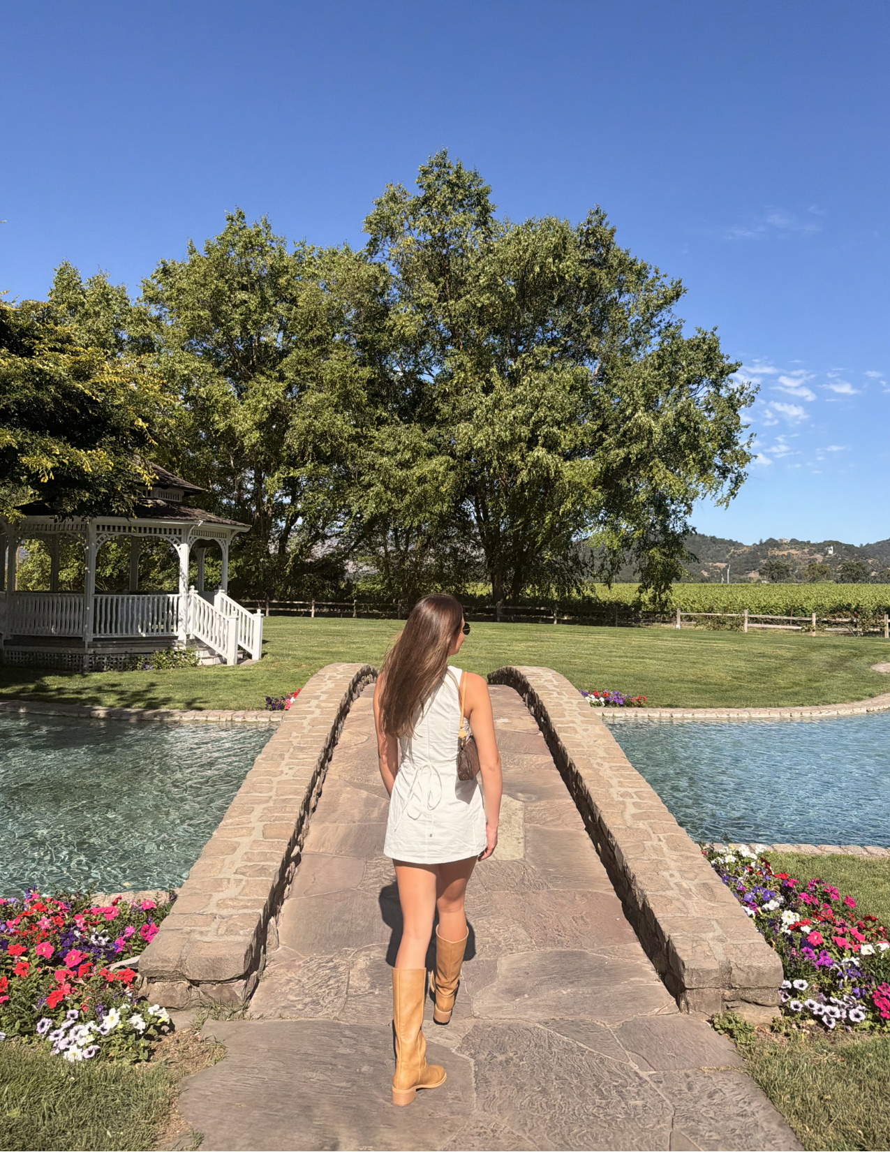 A woman in a white dress and cowboy boots walking across a stone bridge surrounded by colorful flowers and manicured gardens at a beautiful Napa Valley winery estate.
