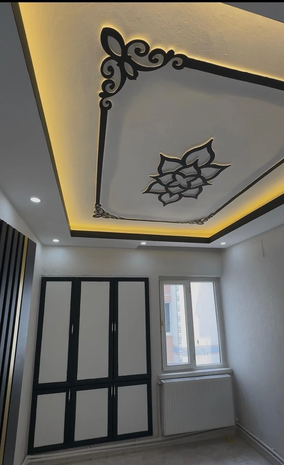 Tavanın dekoratif aydınlatmalı sarkıt ve frizleri, odada doğal ışık alan pencere ve giysi dolabı görünmektedir.