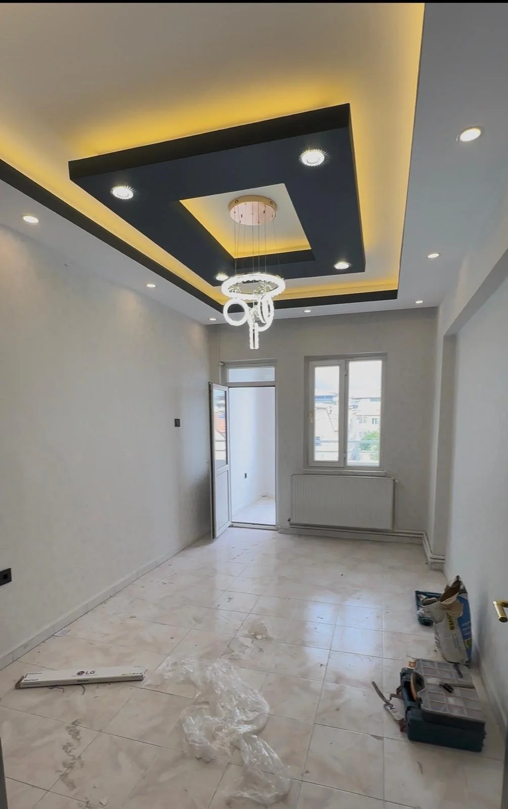 Yeni tamamlanan iç mekan odası, modern tavan tasarımı ve avize ile. Pencere ve kapı açık, yerlerde temizlik malzemeleri ve elektrik aletleri bulunuyor.