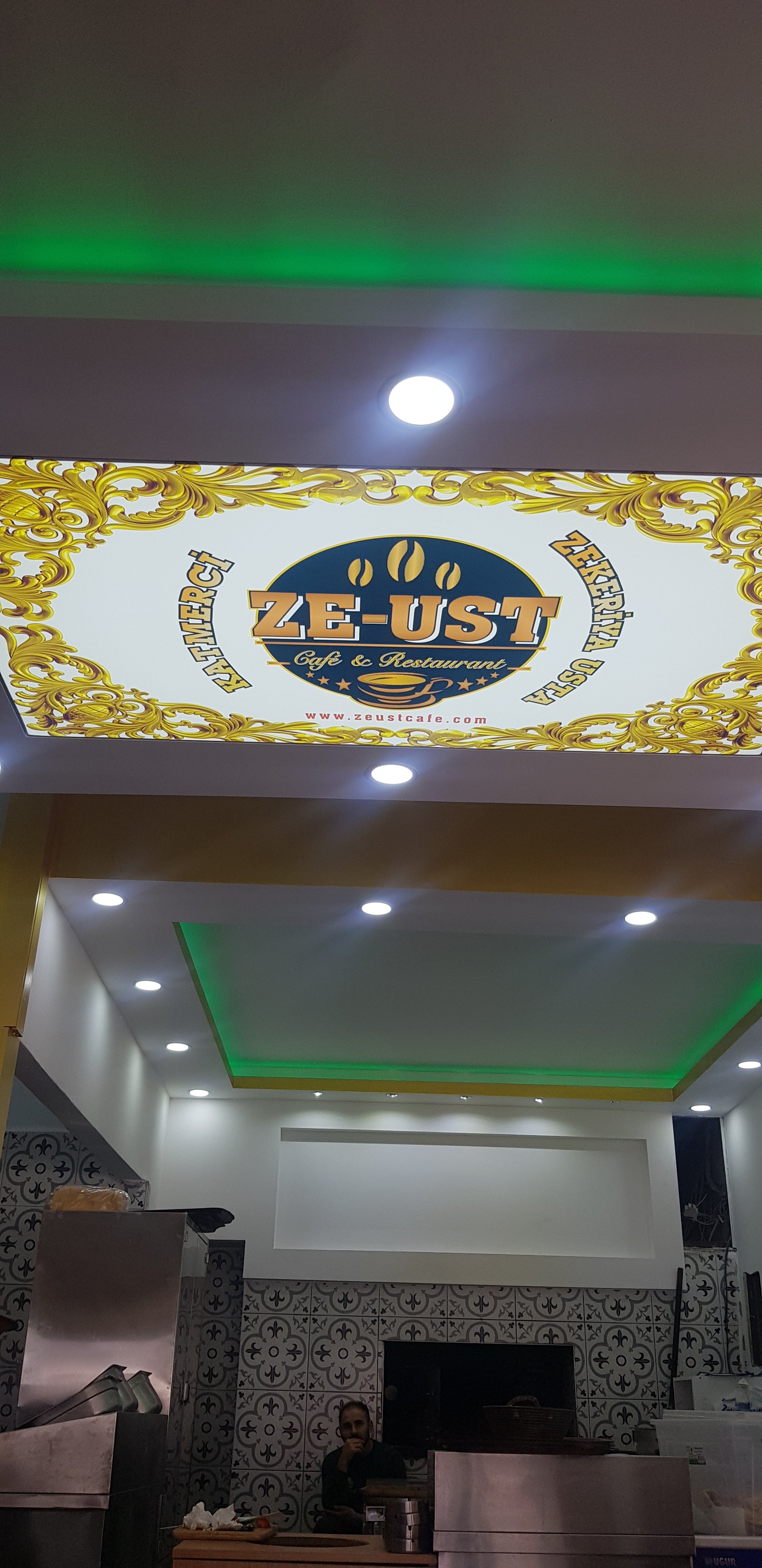 Ze-Ust adlı kafe ve restoranın önündeki ışıklı tabela, altındaki iç mekanda çalışan bir kişi ve dekoratif duvarlar görülüyor.