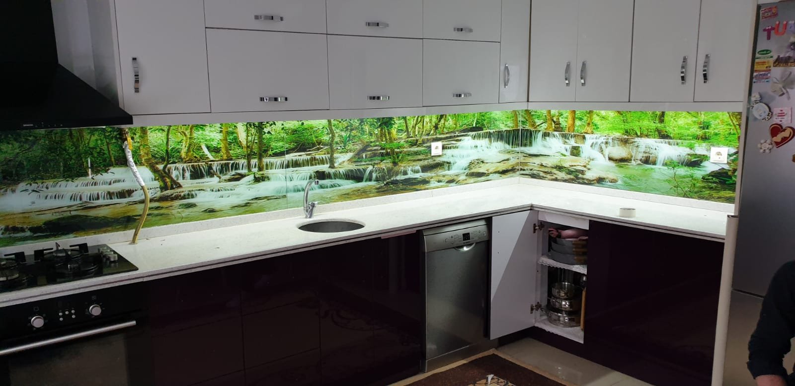 Modern mutfakta yeşil ve şelale manzaralı cam arka plaka ile gömme dolaplar ve küçük lavabo ve gazlı ocak.