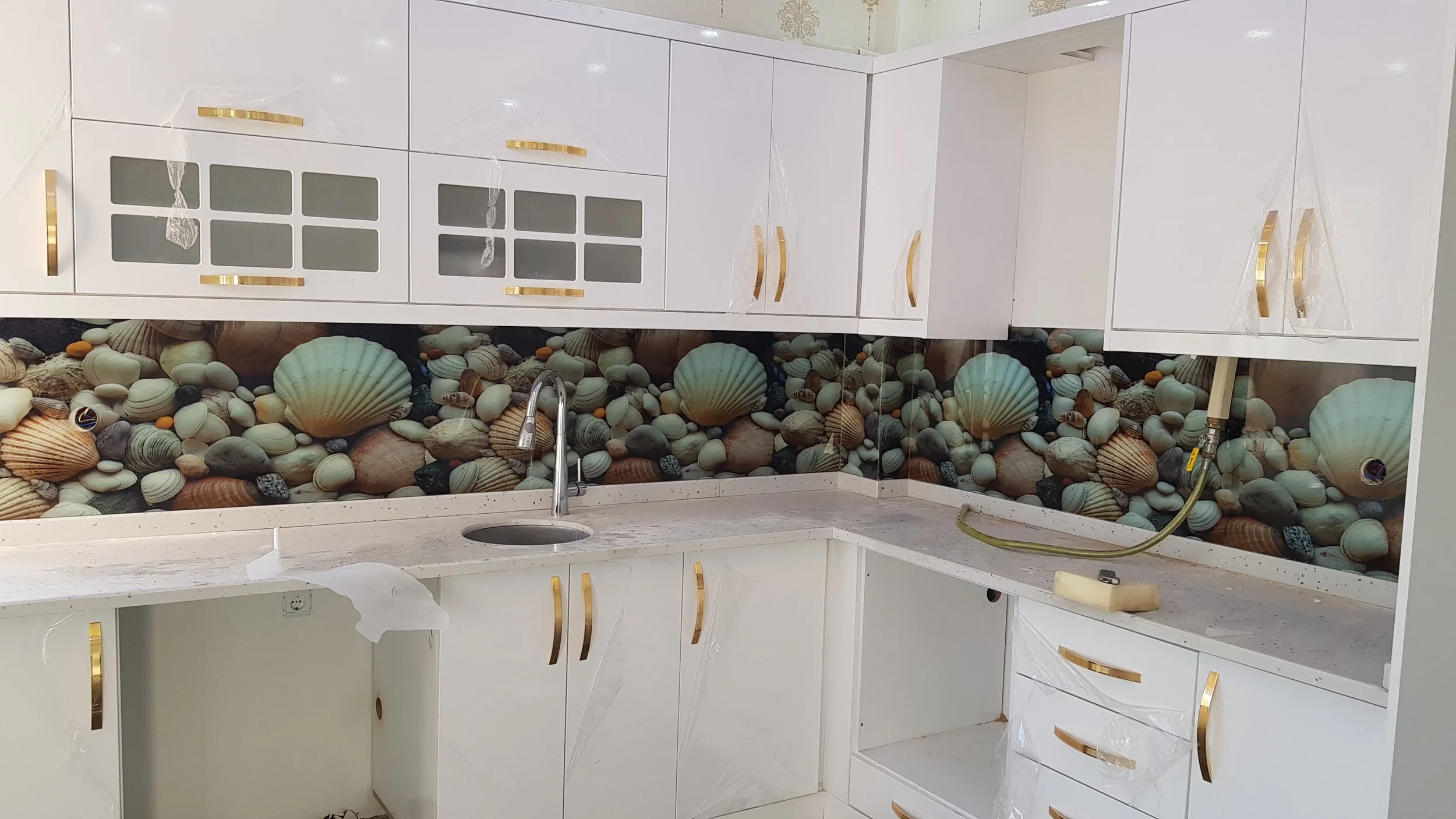 Beyaz dolaplar ve lavabo ile dekoratif deniz kabukları ve taşlar görünüyor. Mutfakta çalışma alanı ve lavabo yer almakta.