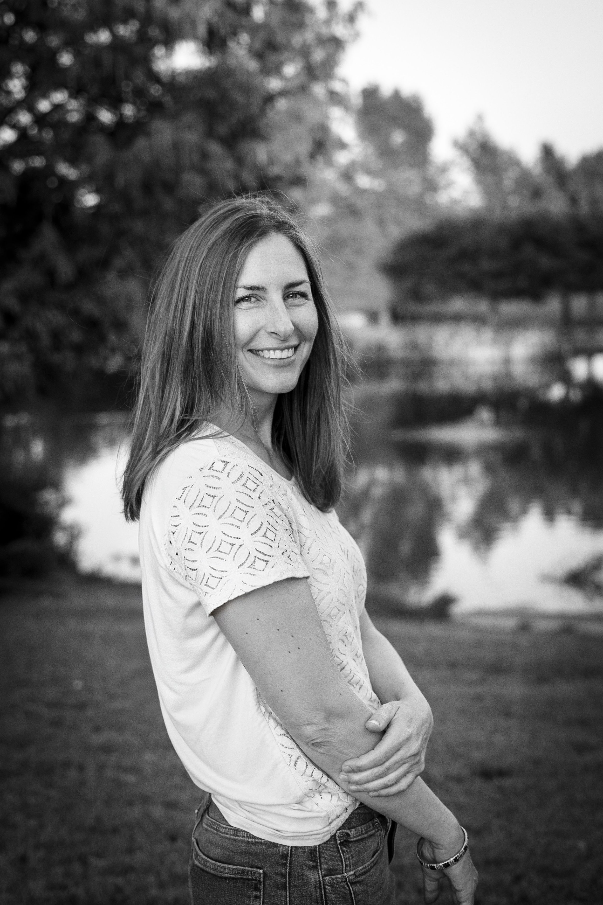 Black and white photo of a woman smiling outdoors. She is wearing a white, short-sleeved, cut-out top and jeans, standing with her arms crossed. The background includes trees and a body of water.
