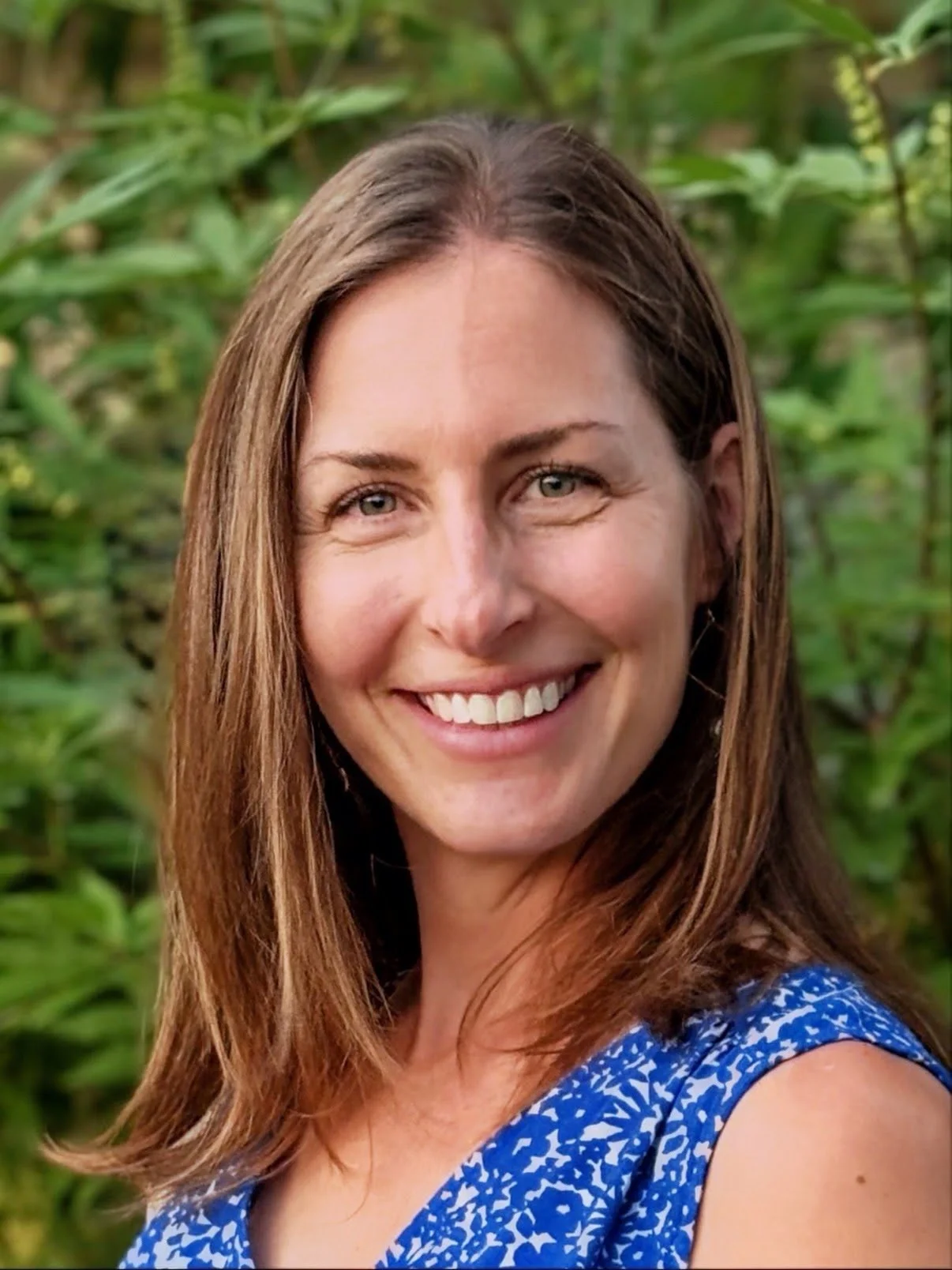 A woman with long brown hair and blue eyes smiling outdoors with green foliage in the background.