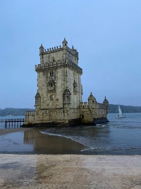 The historic Belem Tower, a stone fortress with battlements and turrets, is situated by the water with a sailboat nearby.