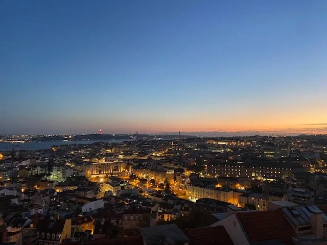 Cityscape during sunset with buildings and streets illuminated, view over a river, and a gradient sky from blue to orange.