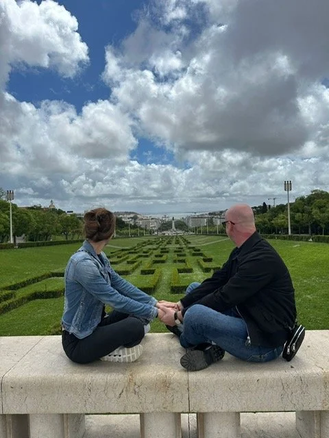 A man and a woman sitting facing each other on a stone ledge in a park, holding hands, with neatly trimmed grass and pathways behind them, under a cloudy sky.